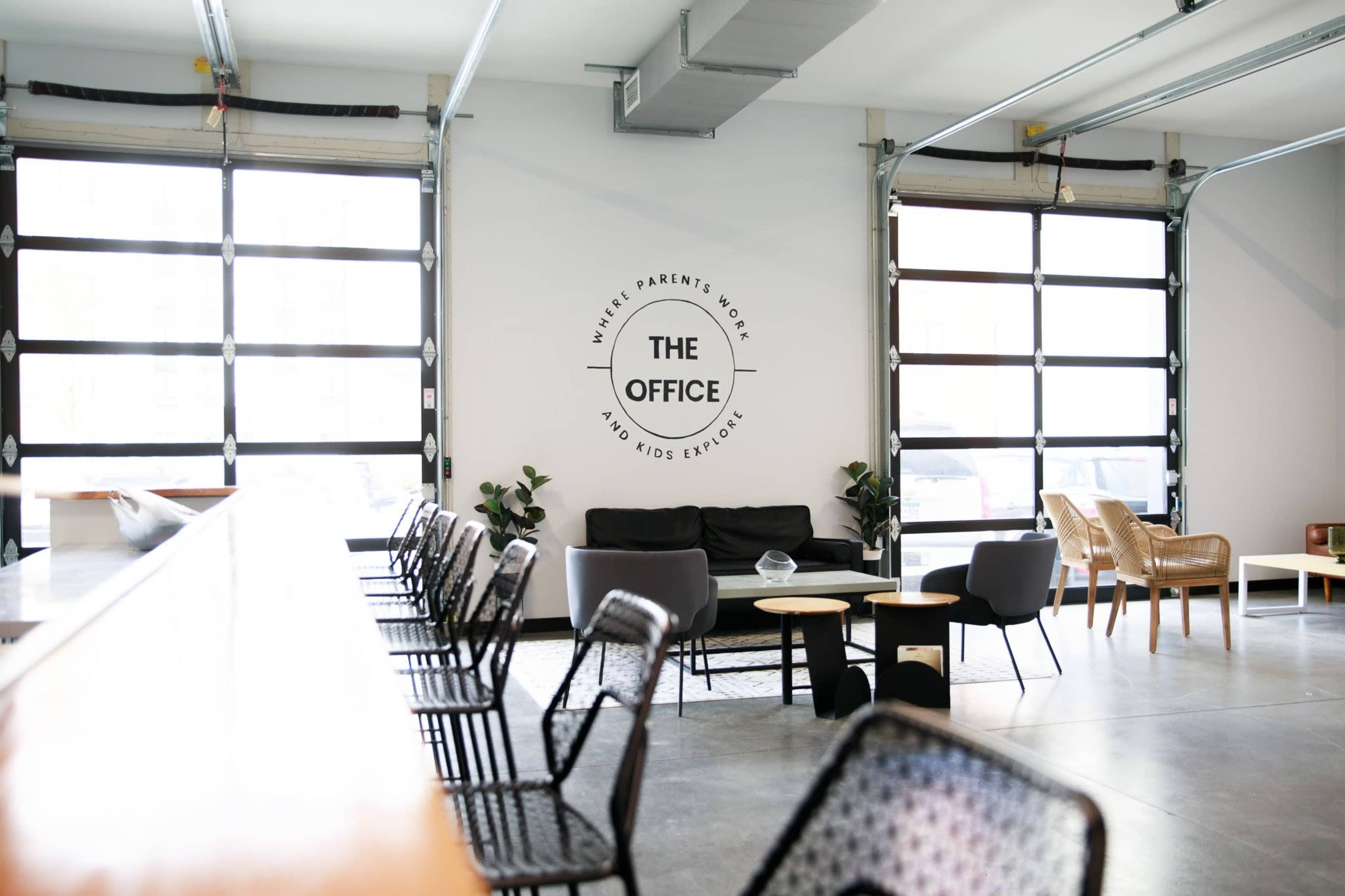 The image shows a modern office space with large windows, a seating area featuring sofas and chairs, and a wall displaying the words "THE OFFICE."