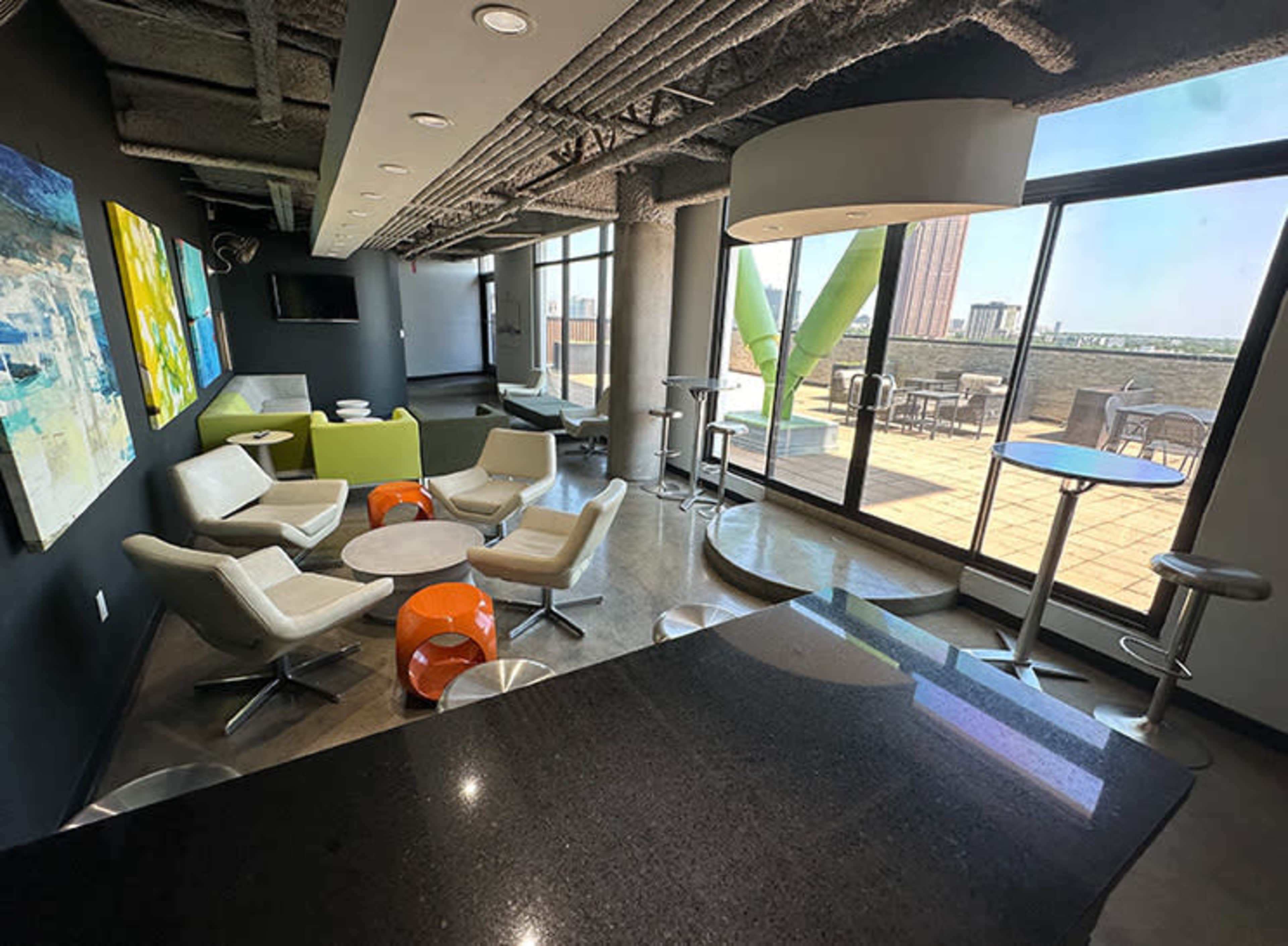 The image shows a modern lounge area with white chairs, orange and white tables, and large windows overlooking an outdoor terrace.