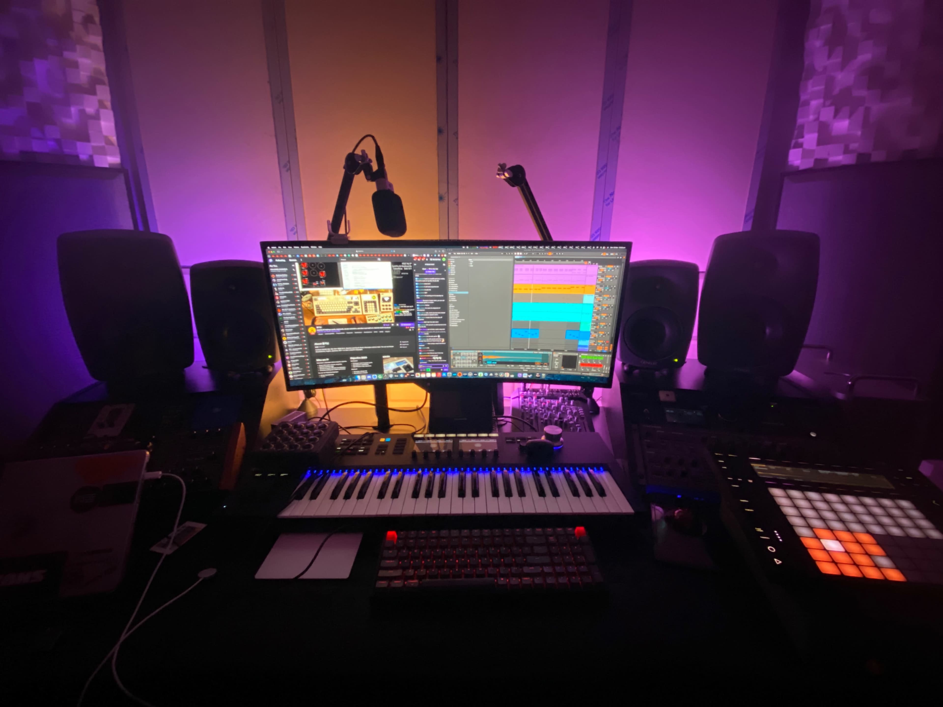 A music production workstation features a keyboard, controllers, a large monitor displaying software, and studio speakers, all illuminated by purple lighting.