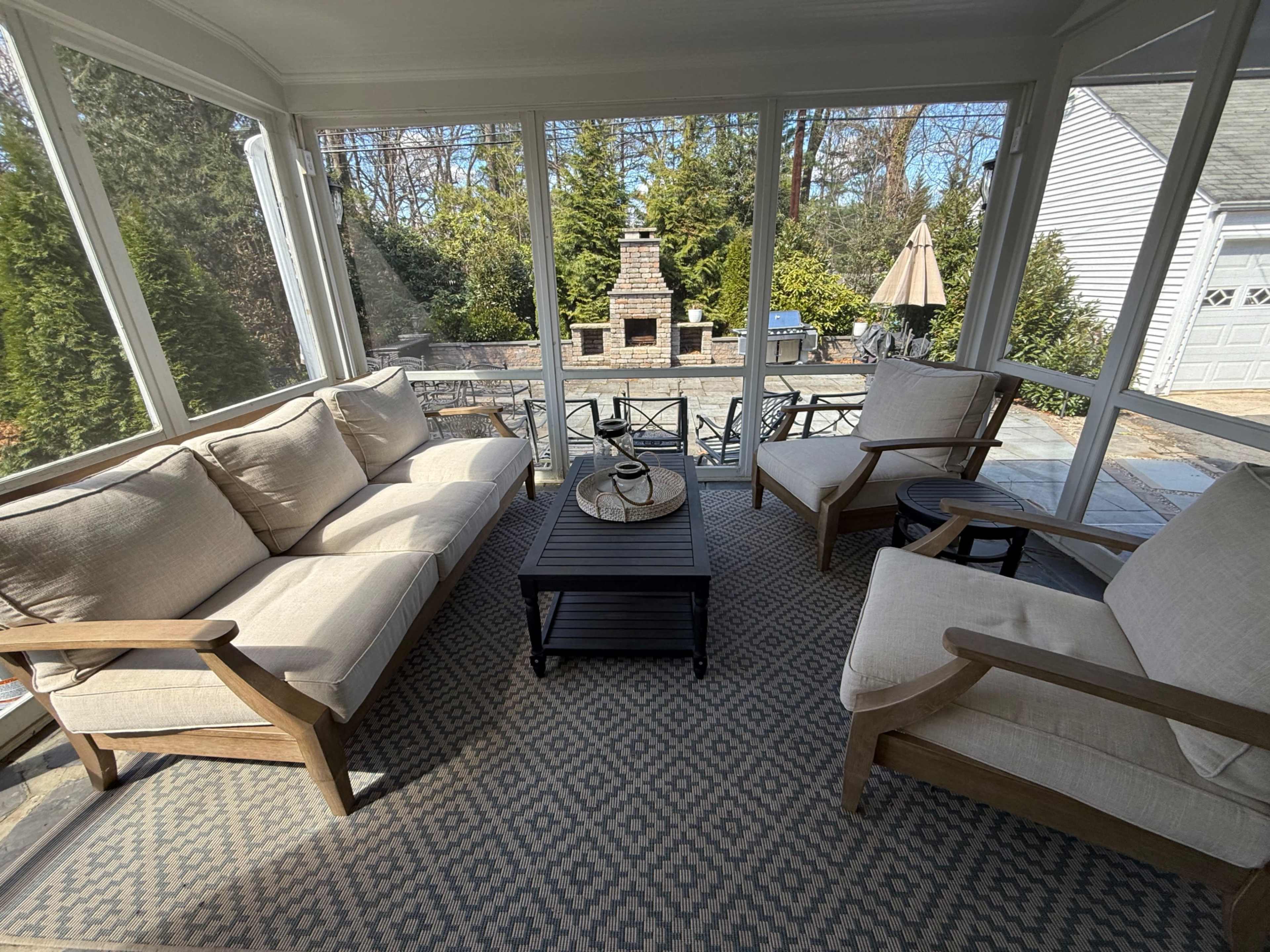 The image shows a screened-in porch with comfortable seating arranged around a central coffee table, overlooking a stone patio and an outdoor fireplace.