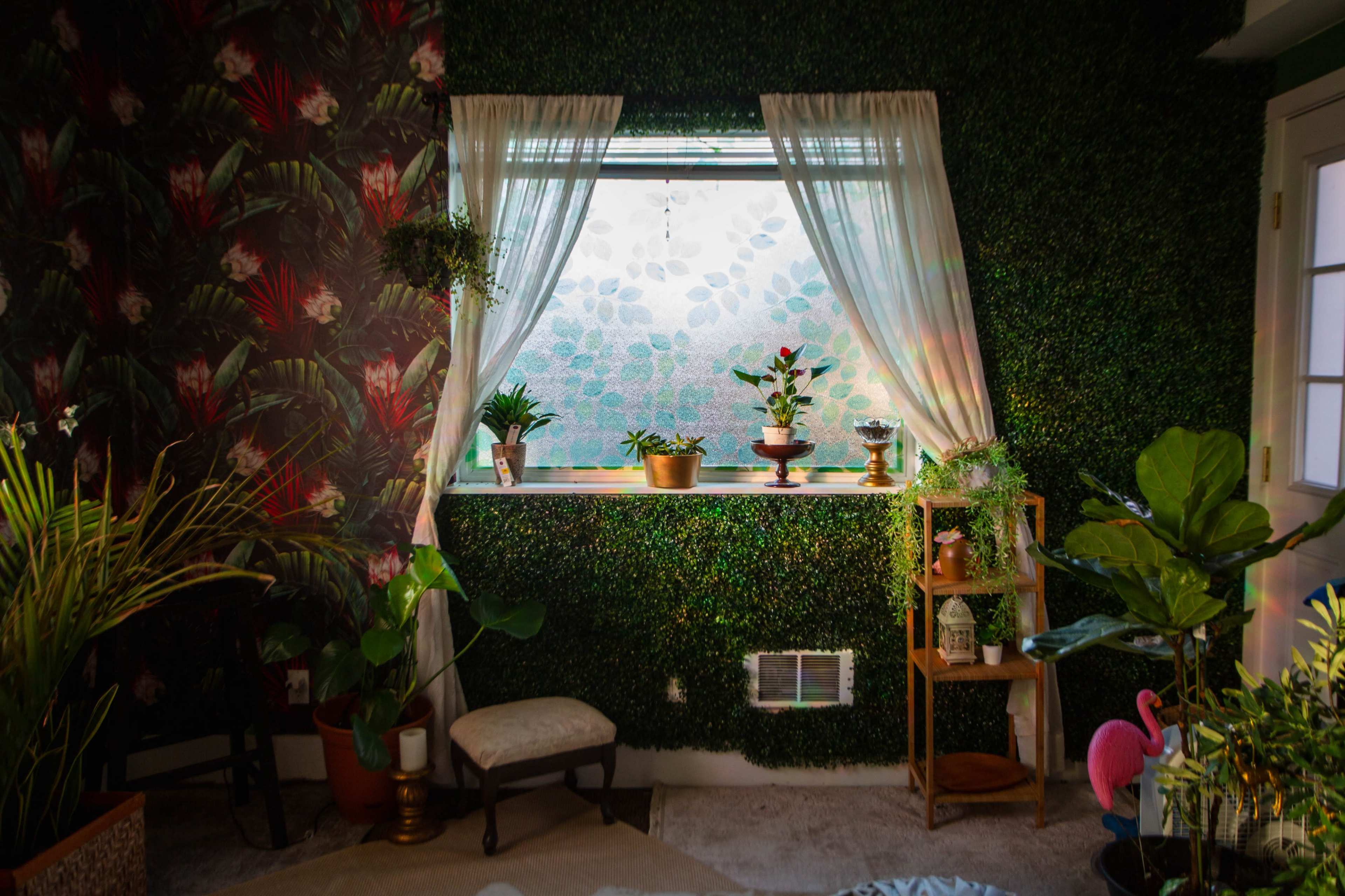 A room with a window framed by sheer curtains, surrounded by green foliage and plants, with floral wallpaper on one wall and a wooden shelf displaying decorative items.