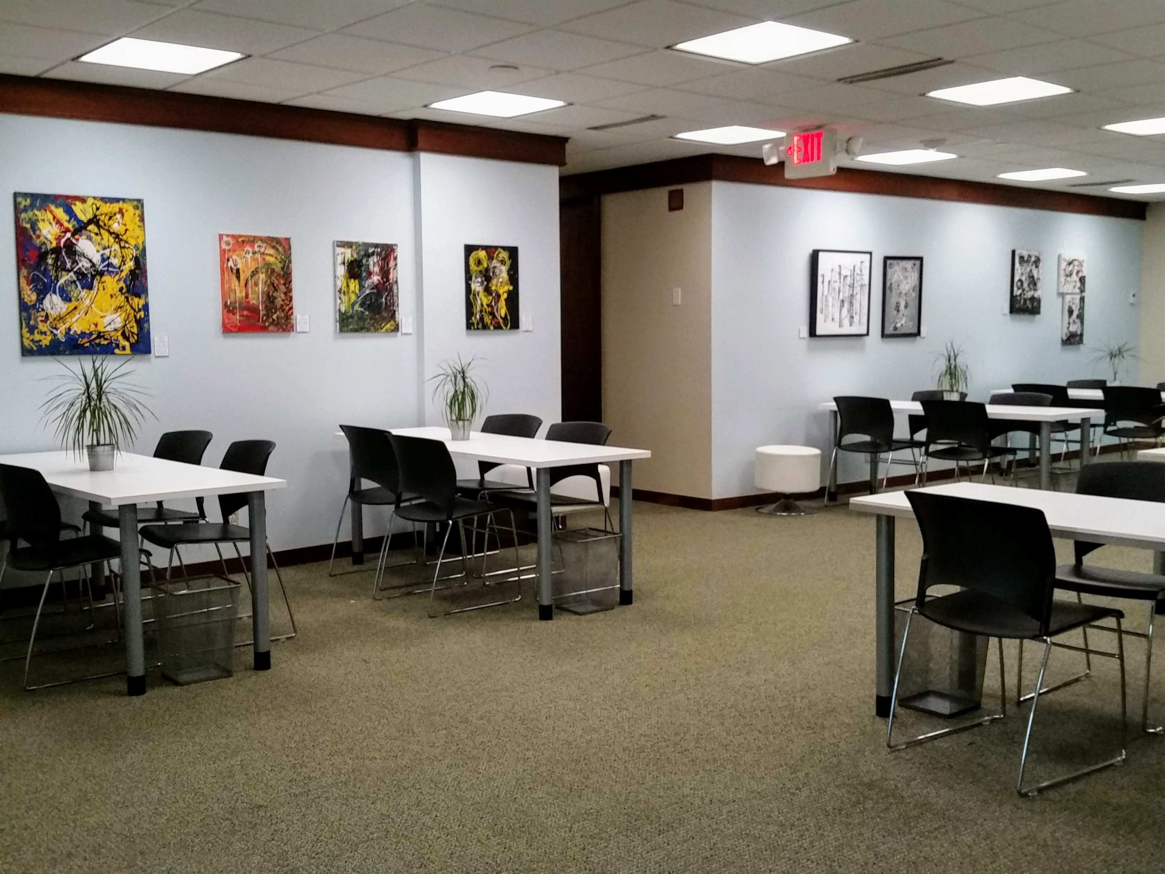 The image shows a spacious room with several tables and chairs, featuring artwork on the walls and plants on the tables.