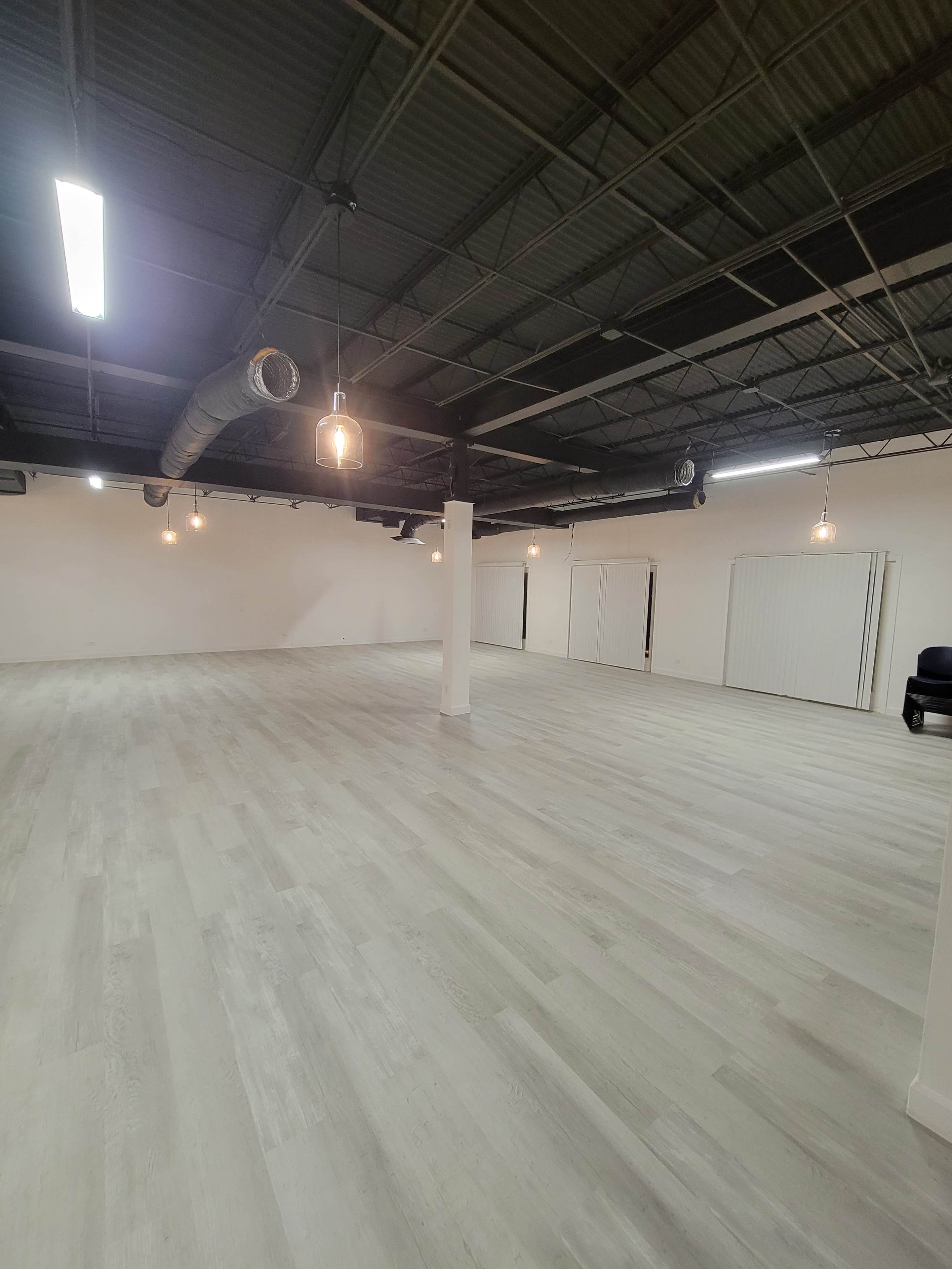 A spacious empty room with light-colored flooring, exposed ductwork, and overhead lighting fixtures.