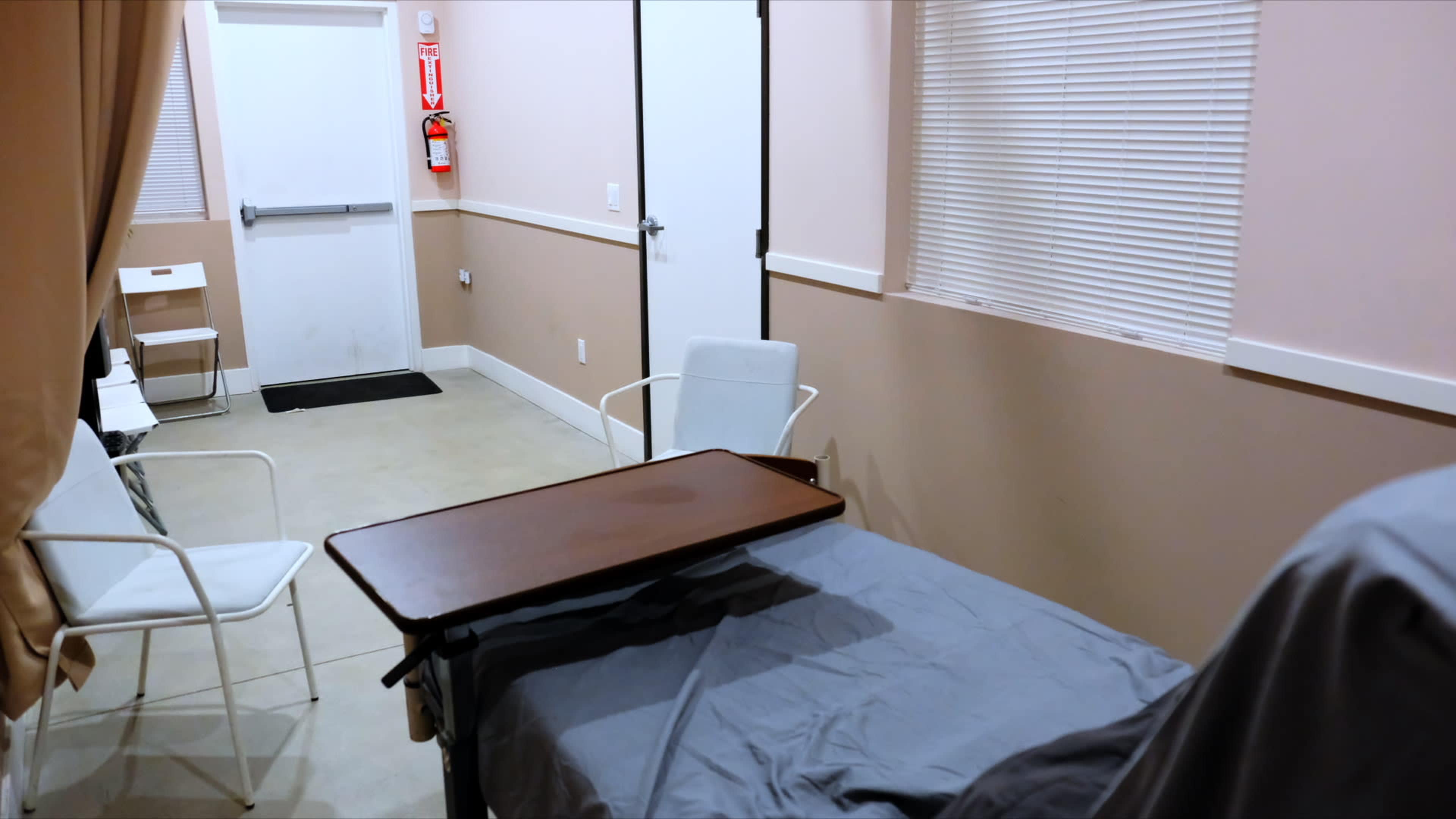 The image shows a simple room with a bed, a table beside it, and two white chairs, along with a door and a window covered by blinds.
