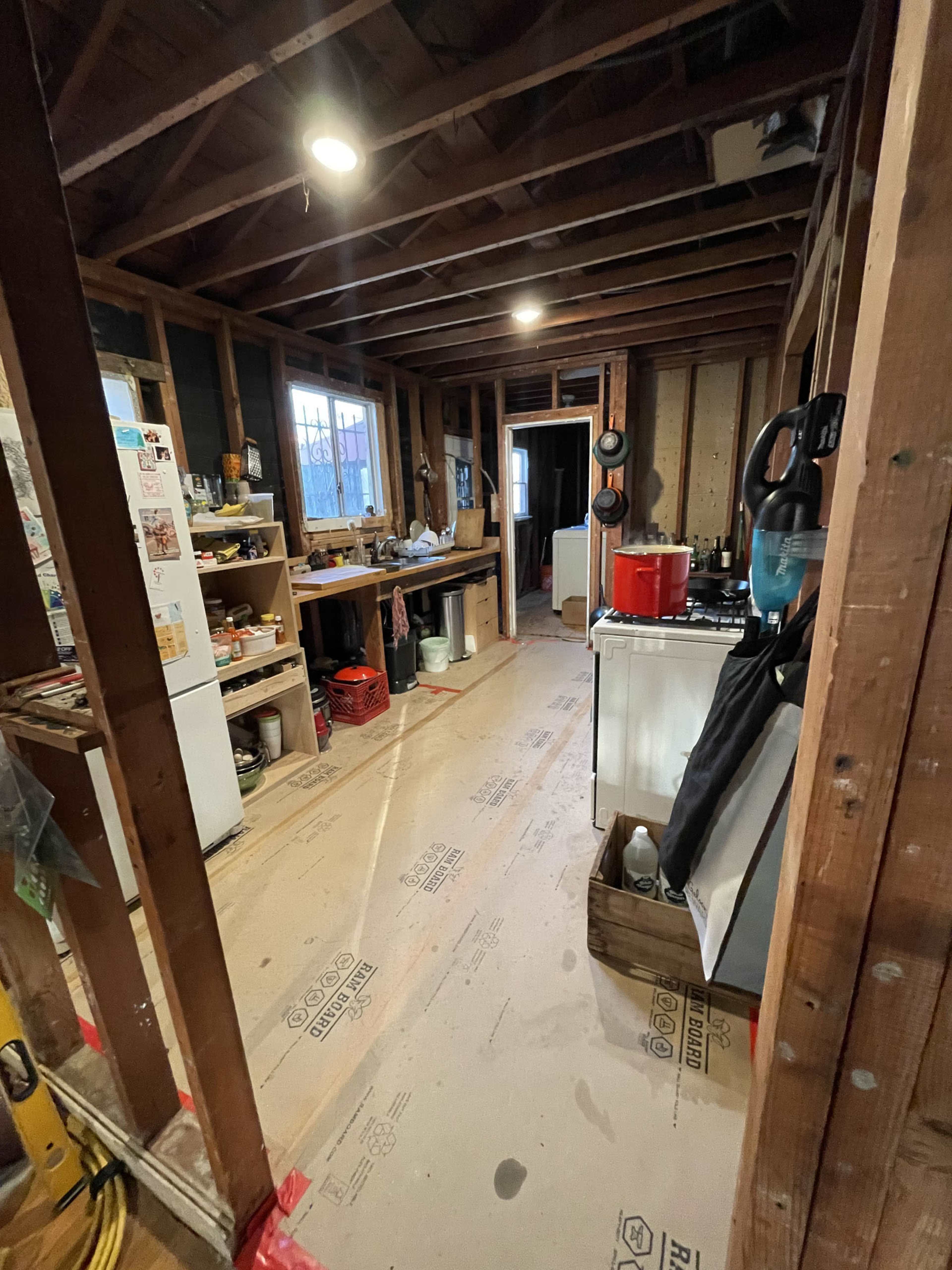 The image shows an unfinished room with exposed wooden beams, bare walls, and various items arranged on shelves along one side.