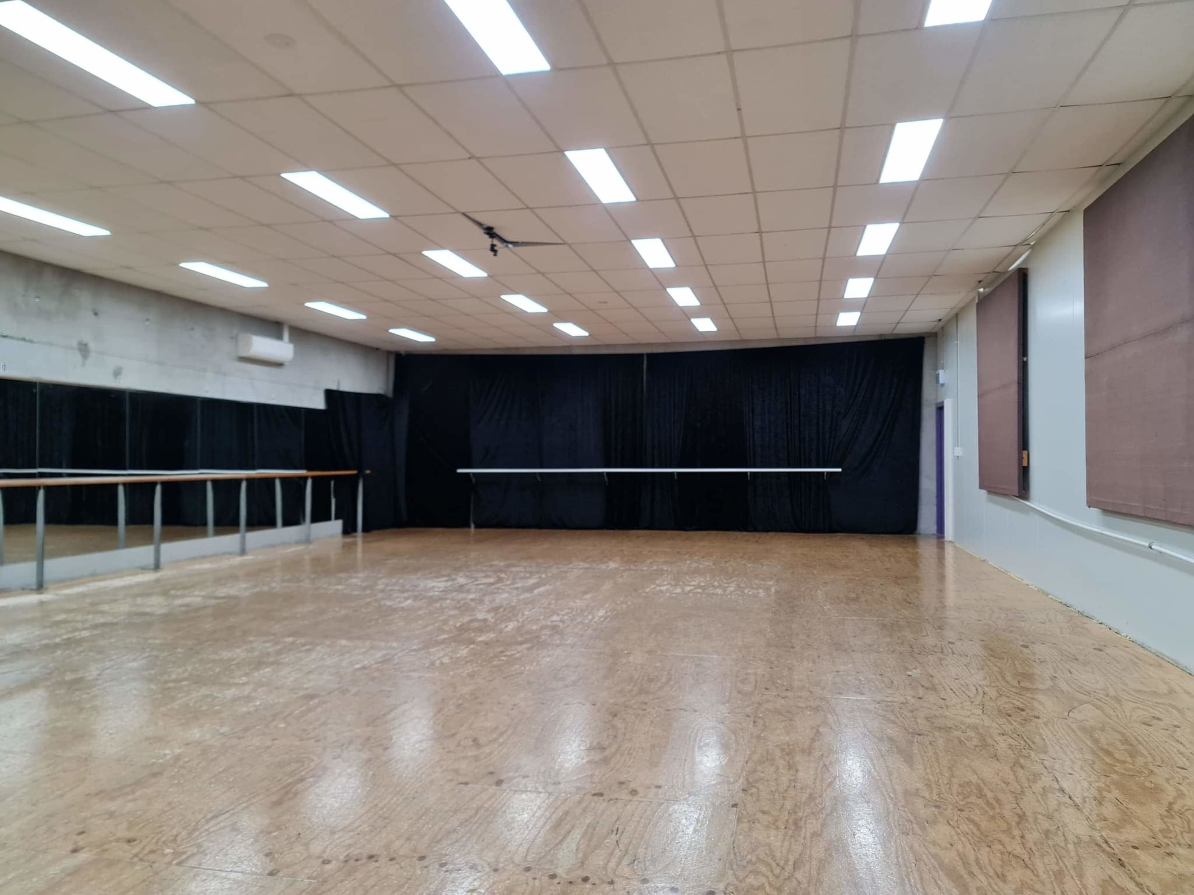 A spacious dance studio with a wooden floor, mirrors on one wall, and black curtains along another.