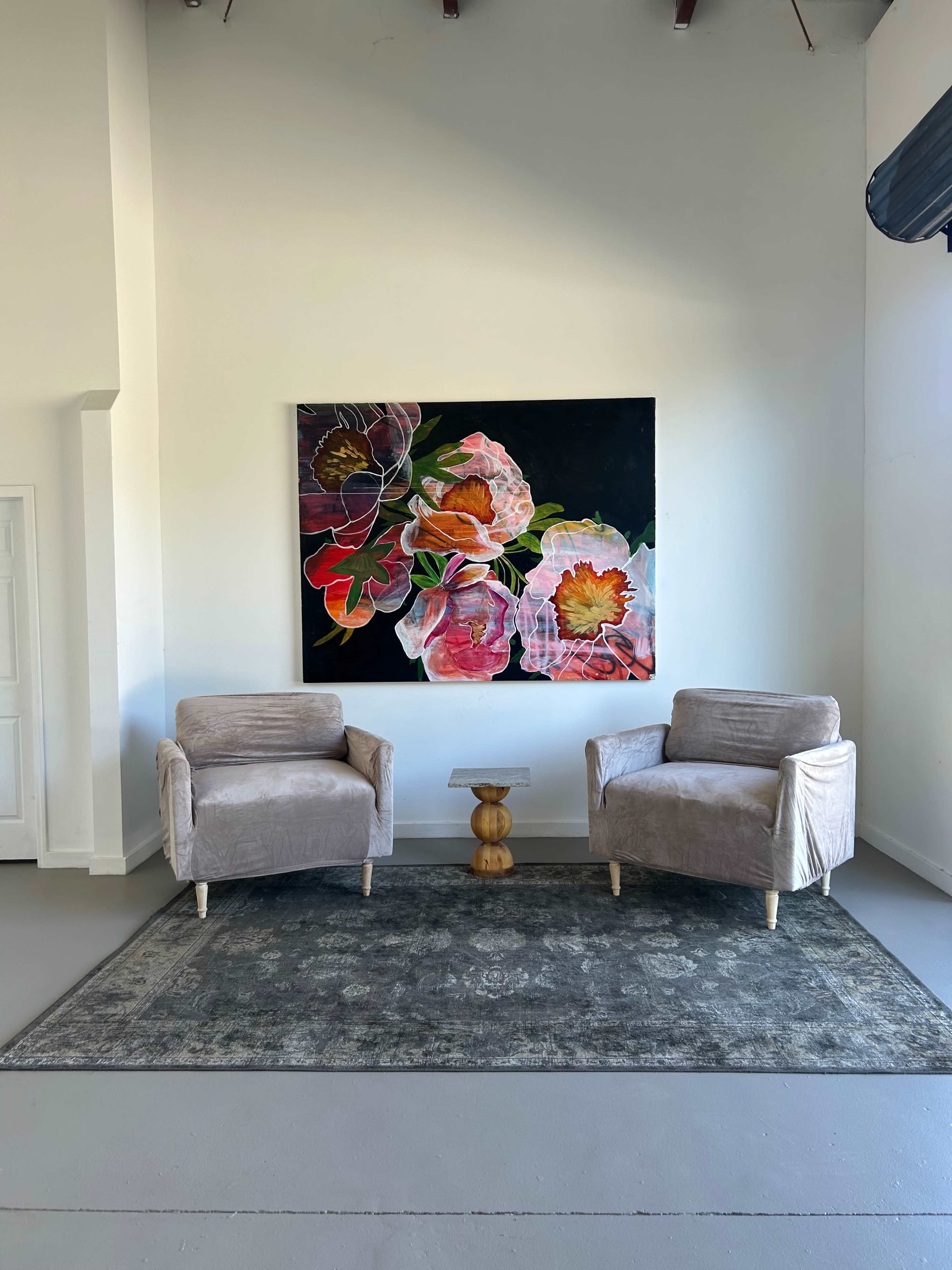 The image shows two light-colored armchairs facing a small wooden side table, situated on a rug in a room with a colorful floral painting on the wall.