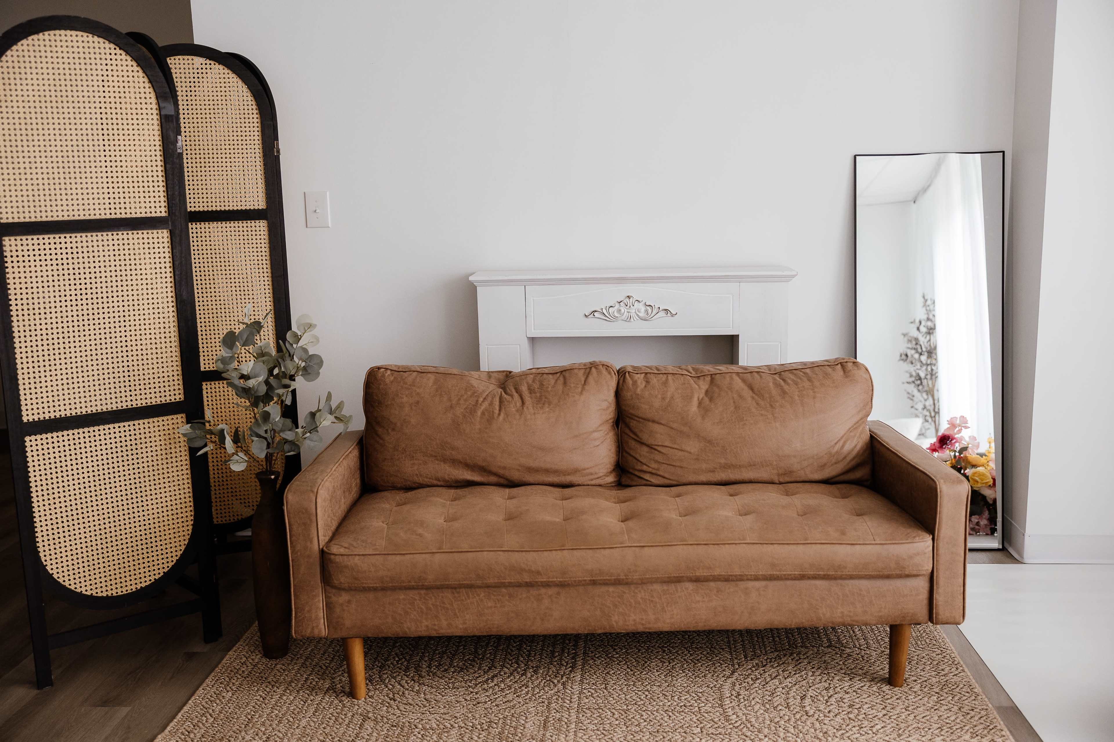 The image shows a brown fabric couch placed in a room with a wooden screen, an empty white mantel, and a tall mirror.