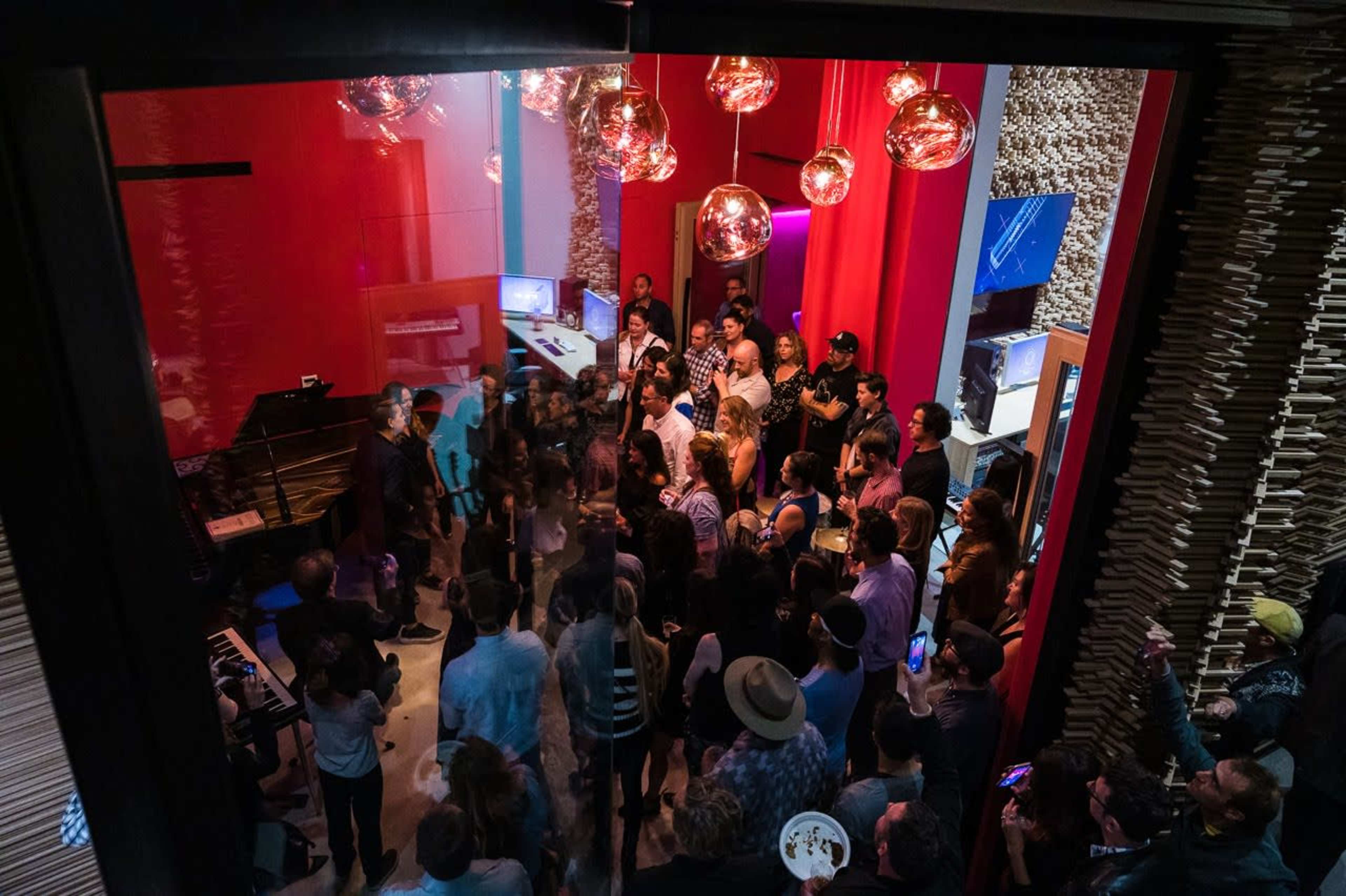 A crowded indoor space features a group of people gathered around a performer while colorful lighting illuminates the room.