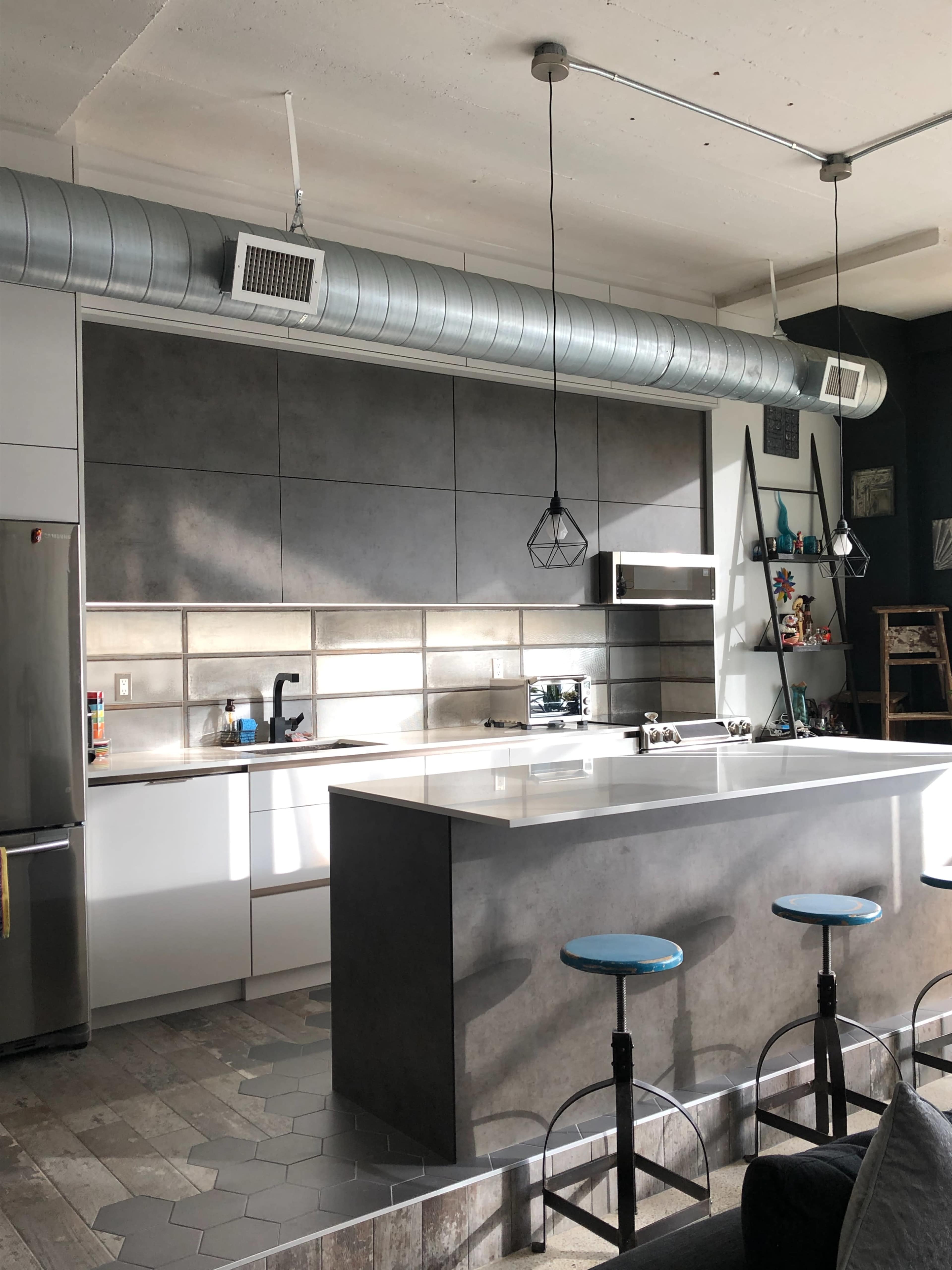 The image shows a modern kitchen with a gray and white color scheme, featuring an island with blue stools and industrial-style lighting.
