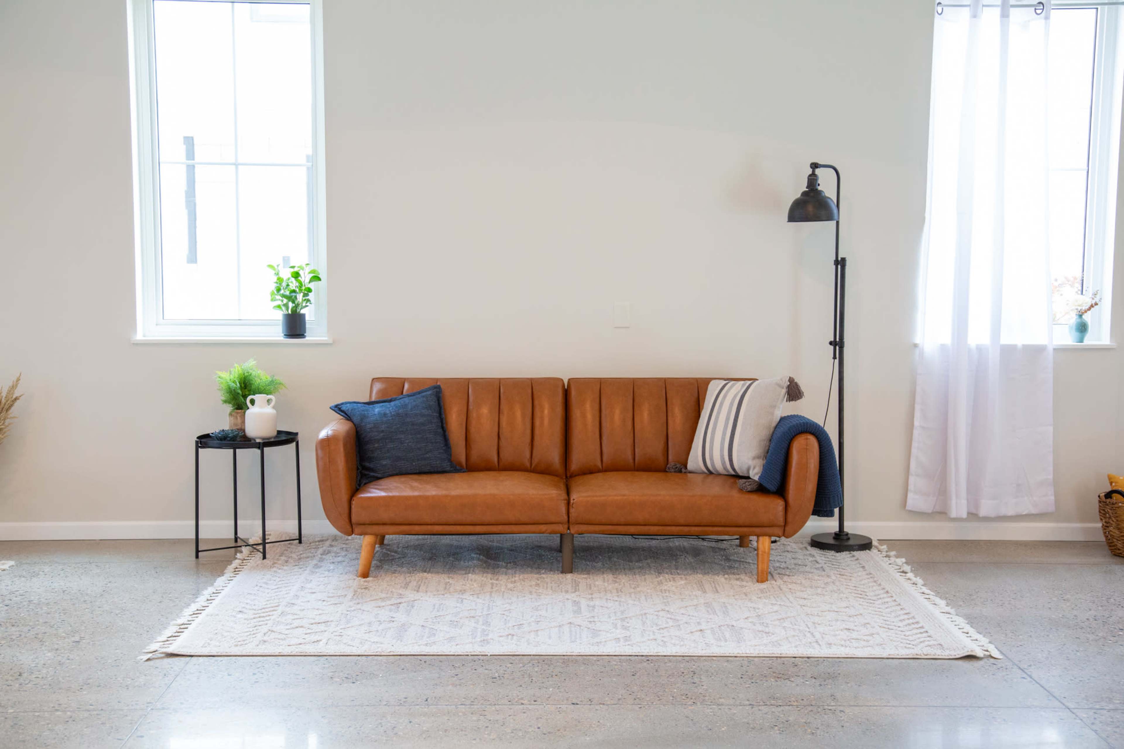 A brown leather sofa is positioned against a light-colored wall, accompanied by a small side table and a floor lamp with a white curtain in the background.
