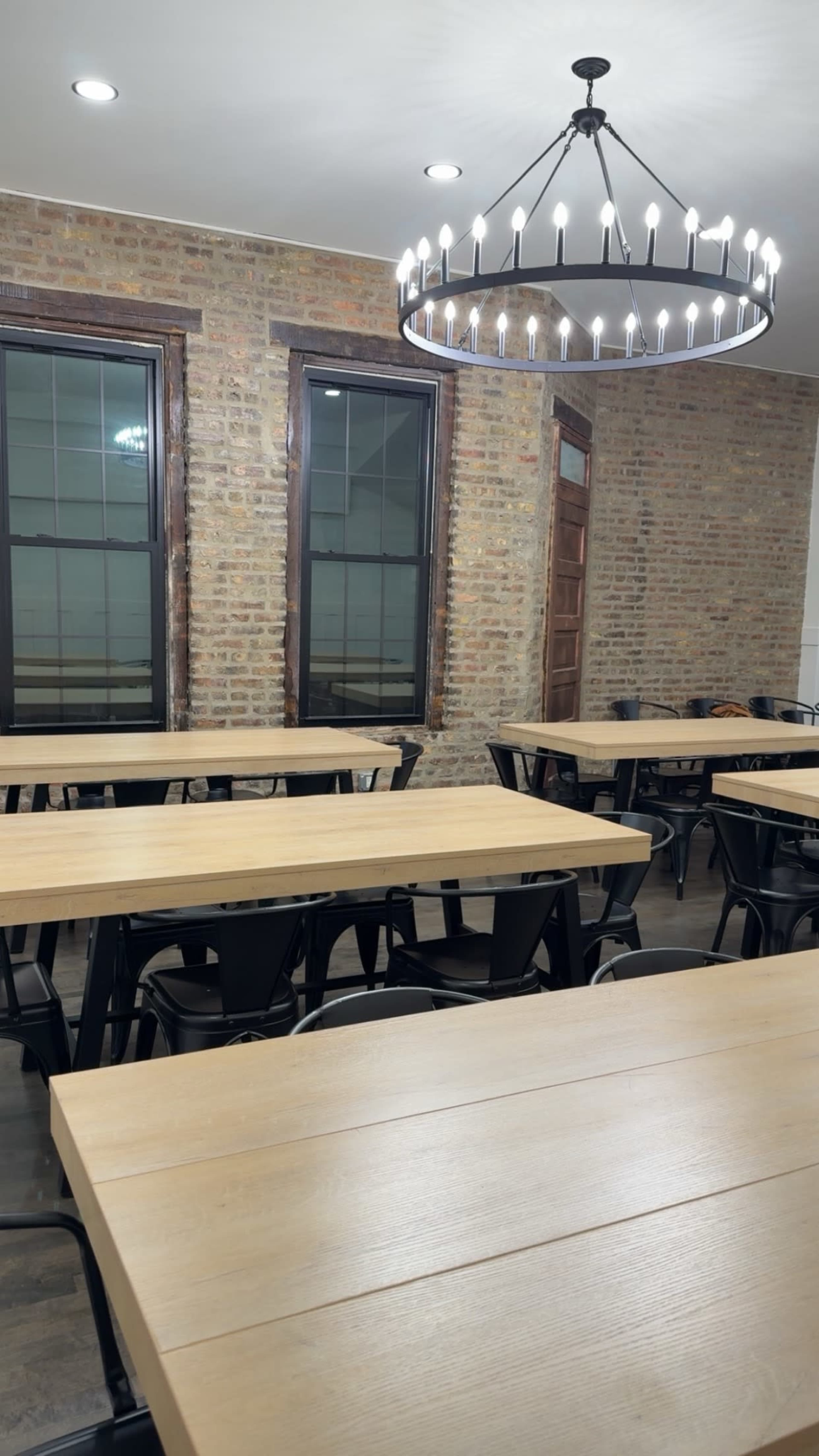 The image shows a spacious classroom with several wooden tables arranged in rows, exposed brick walls, and large windows letting in light from outside.