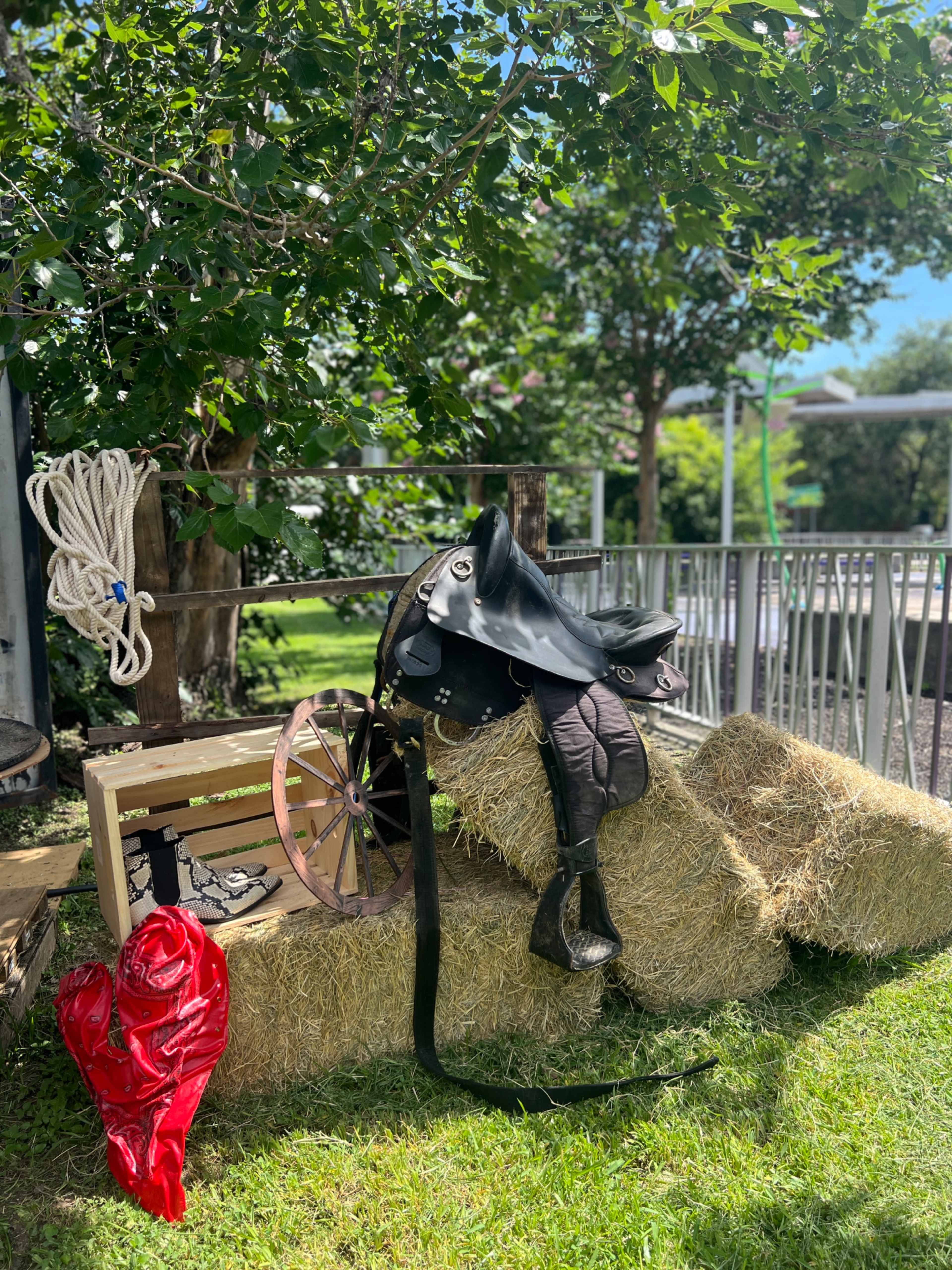 A saddle, lasso, and a wooden cart wheel rest on bales of hay under a tree.
