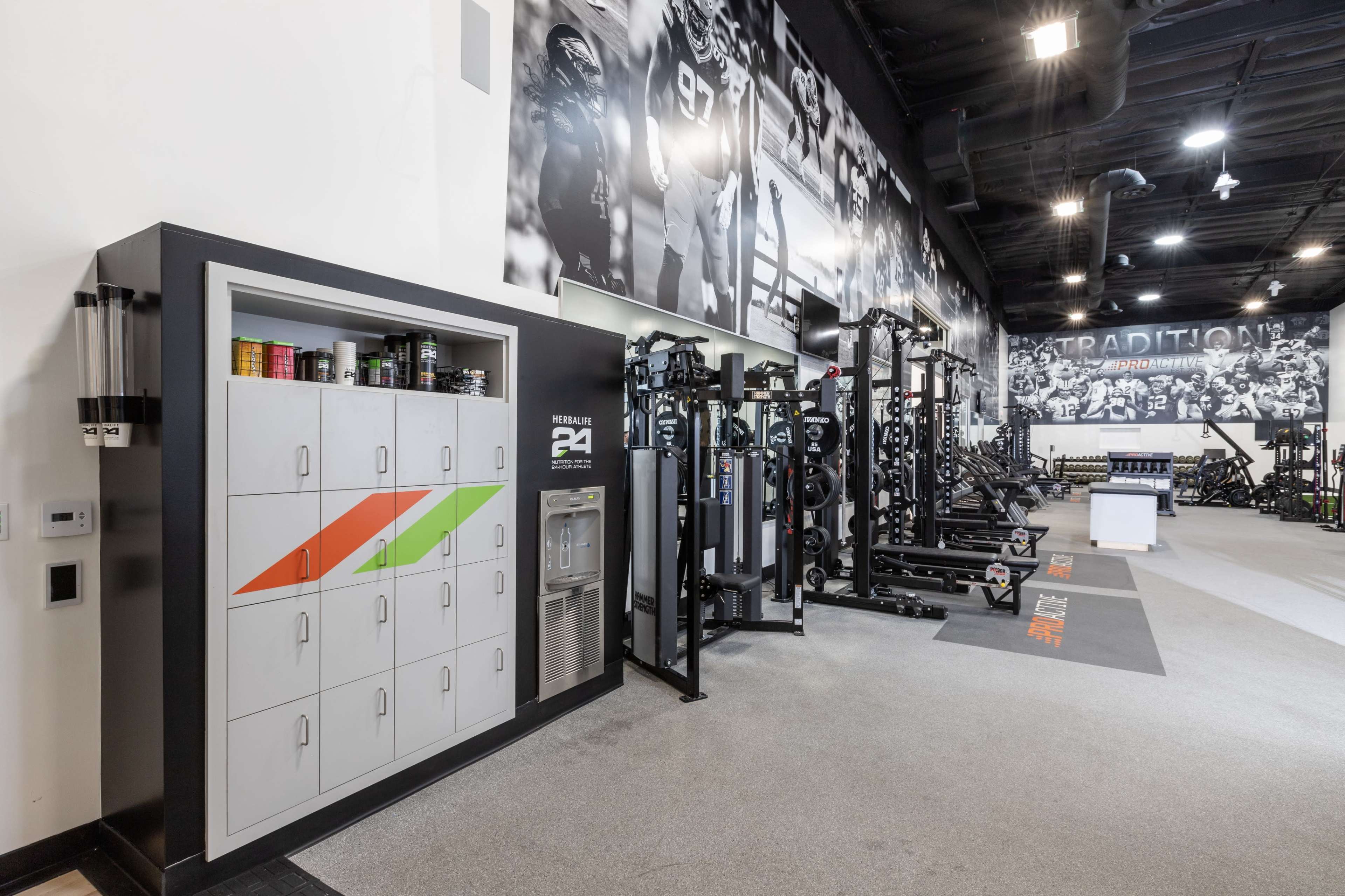 The image shows a modern gym interior featuring workout equipment, locker storage, and a wall adorned with large black and white graphics.