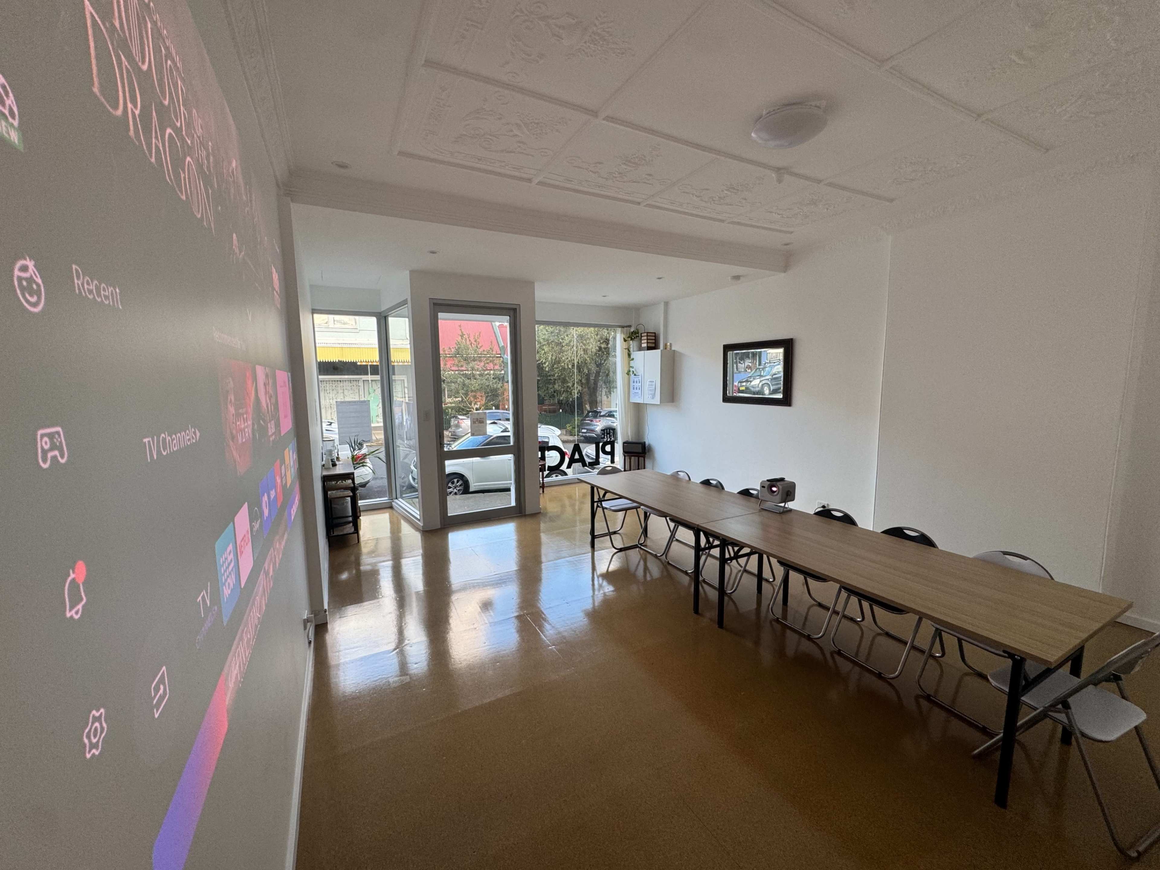 The image shows a spacious room with a large projection screen displaying a menu, a long wooden table with chairs, and a glass entryway that leads to an exterior view of a street.