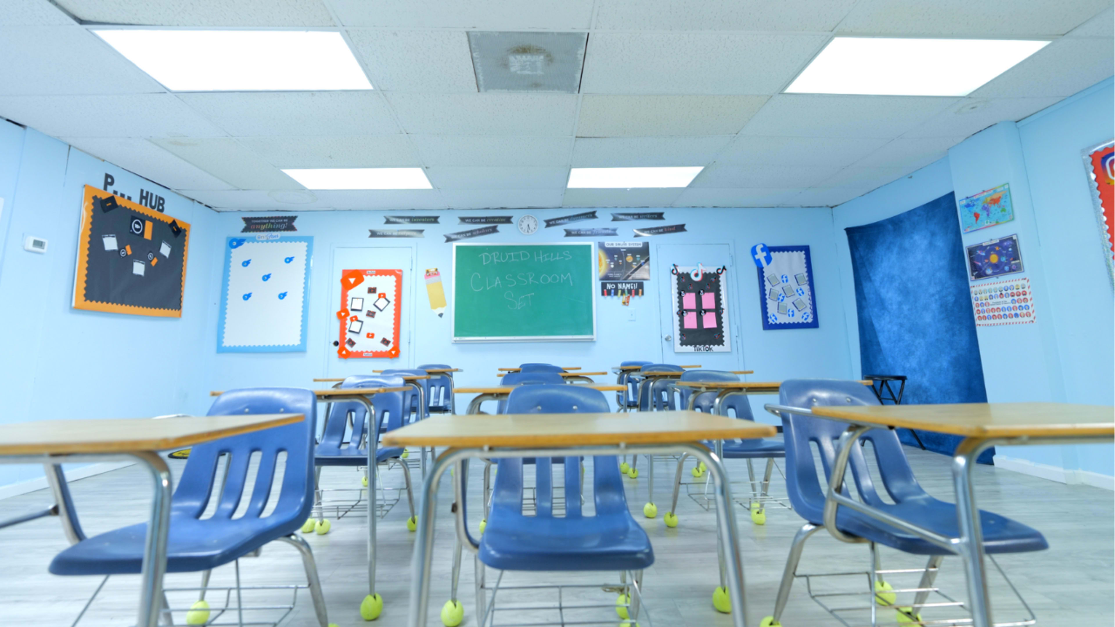 The image shows a classroom with several empty blue desks, a chalkboard at the front, and colorful bulletin boards on the walls.