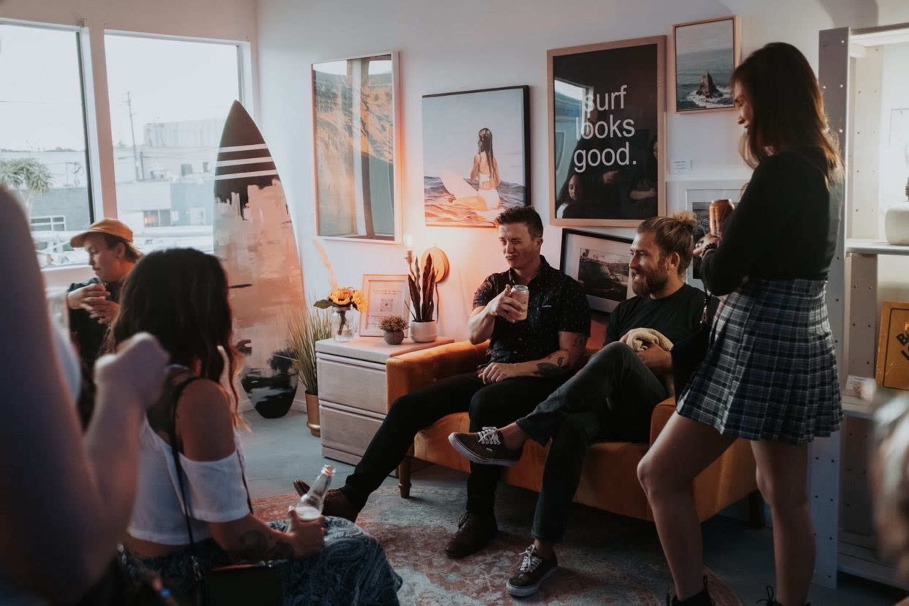 A group of people socialize inside a room decorated with artwork and a surfboard, while seated on an orange couch and holding drinks.