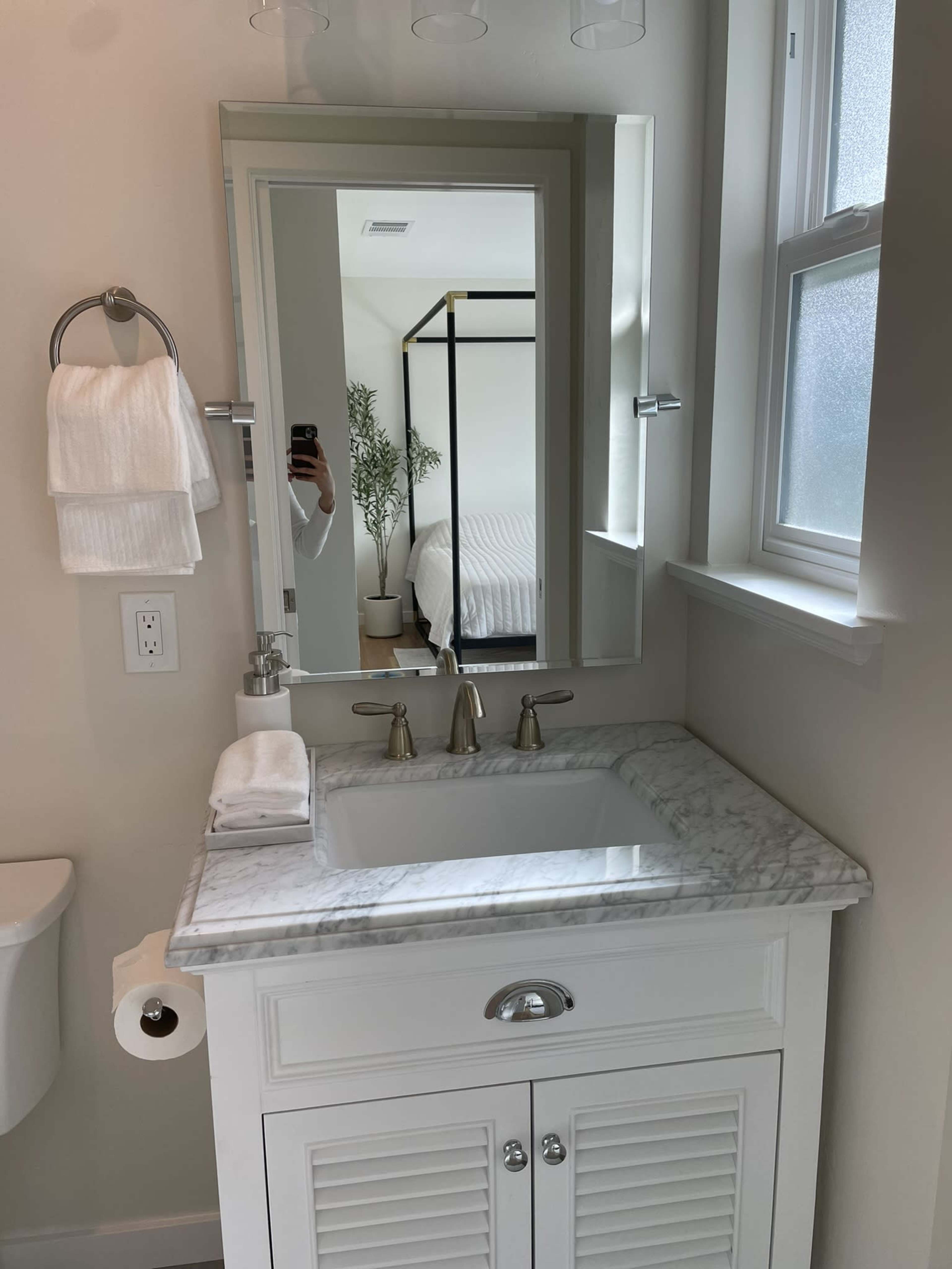 A white bathroom features a marble countertop with a sink, a large mirror, and a towel rack, with a glimpse of a bedroom visible in the background.