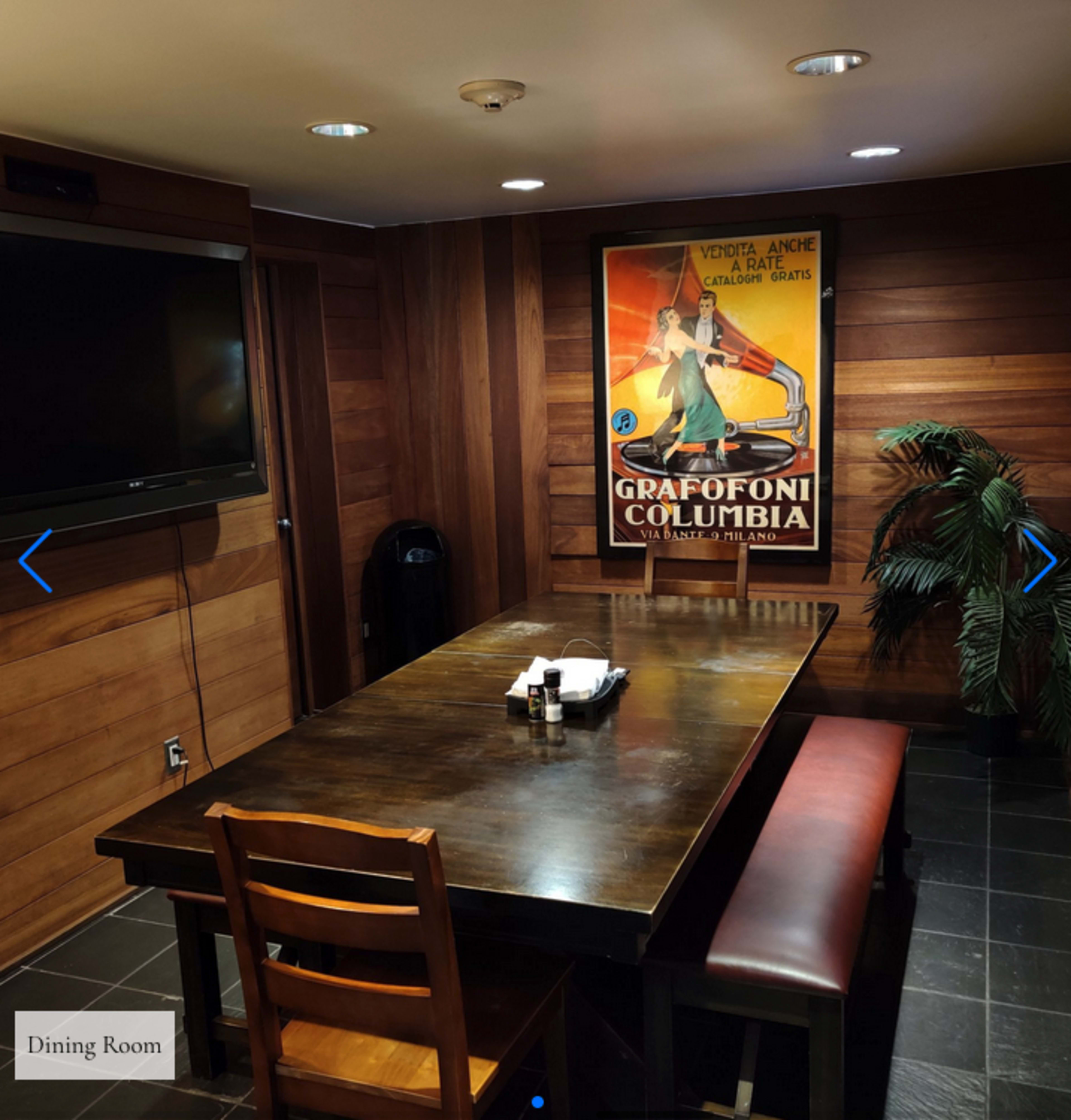 A dining room featuring a large wooden table, a chair, a retro poster on the wall, and a small plant in the corner.