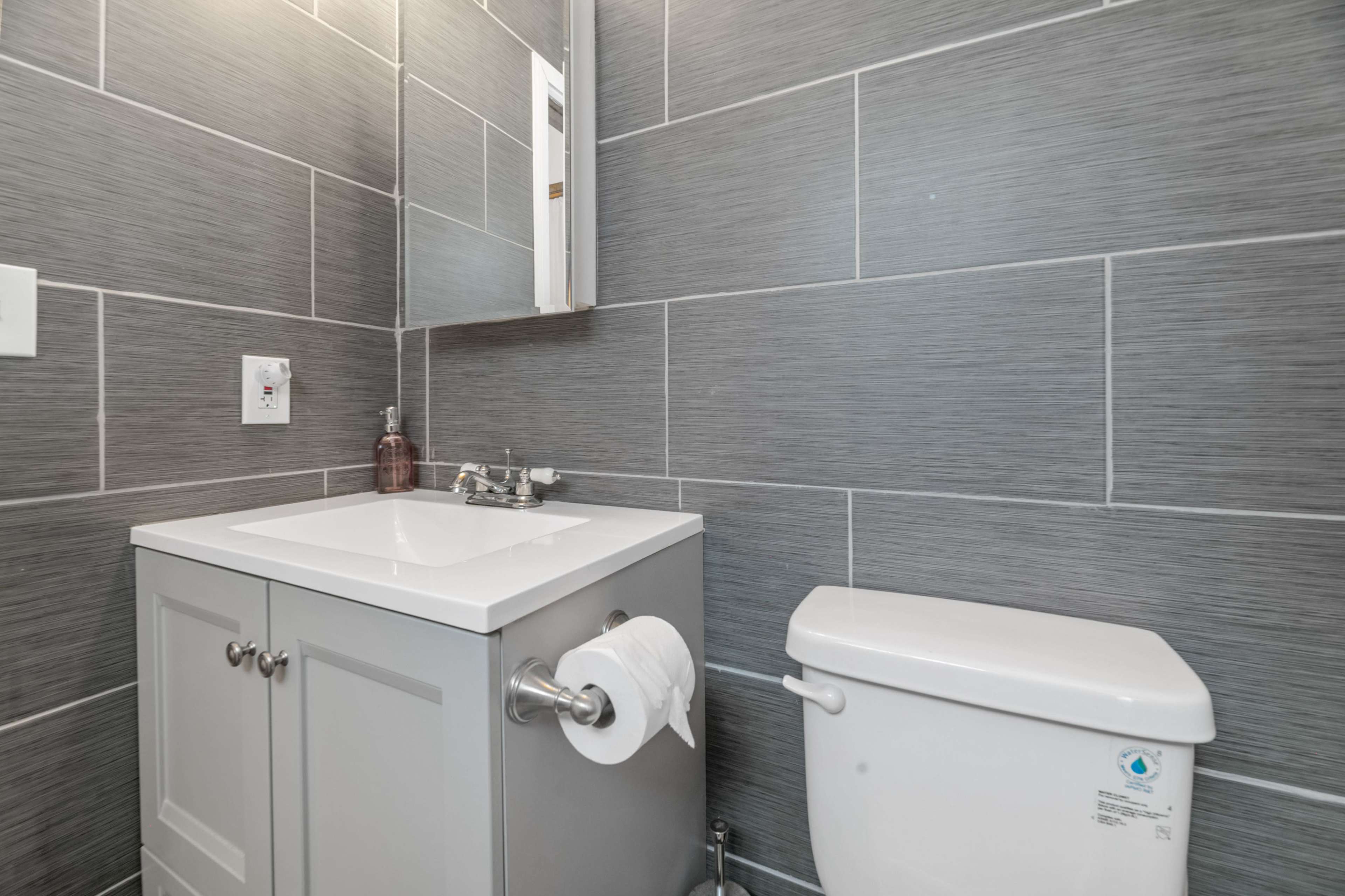 A modern bathroom featuring a gray tiled wall, a white sink cabinet with a mirror above, and a white toilet beside it.