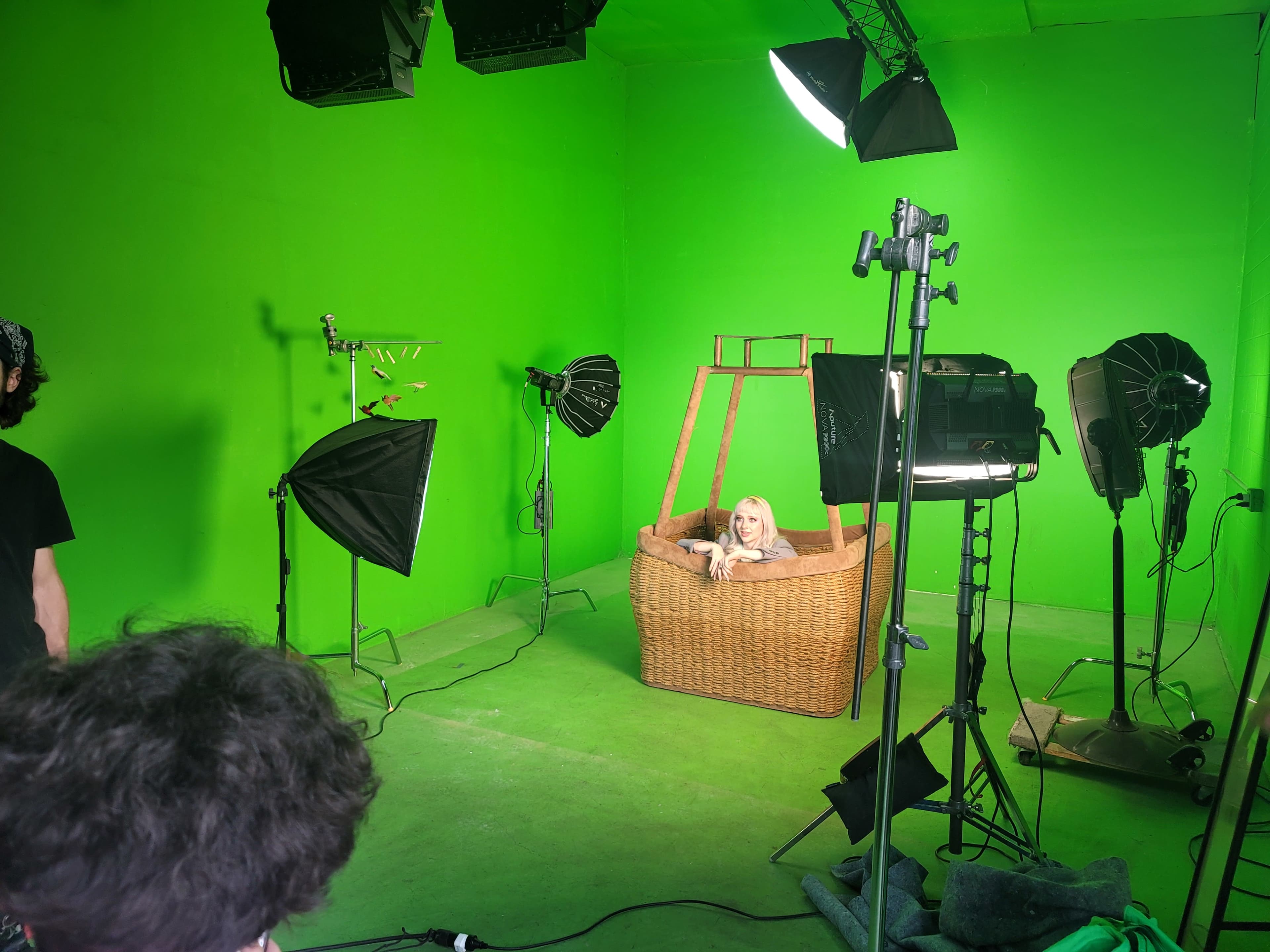 The scene shows a model sitting in a wicker basket surrounded by studio lighting in a green-screen room.
