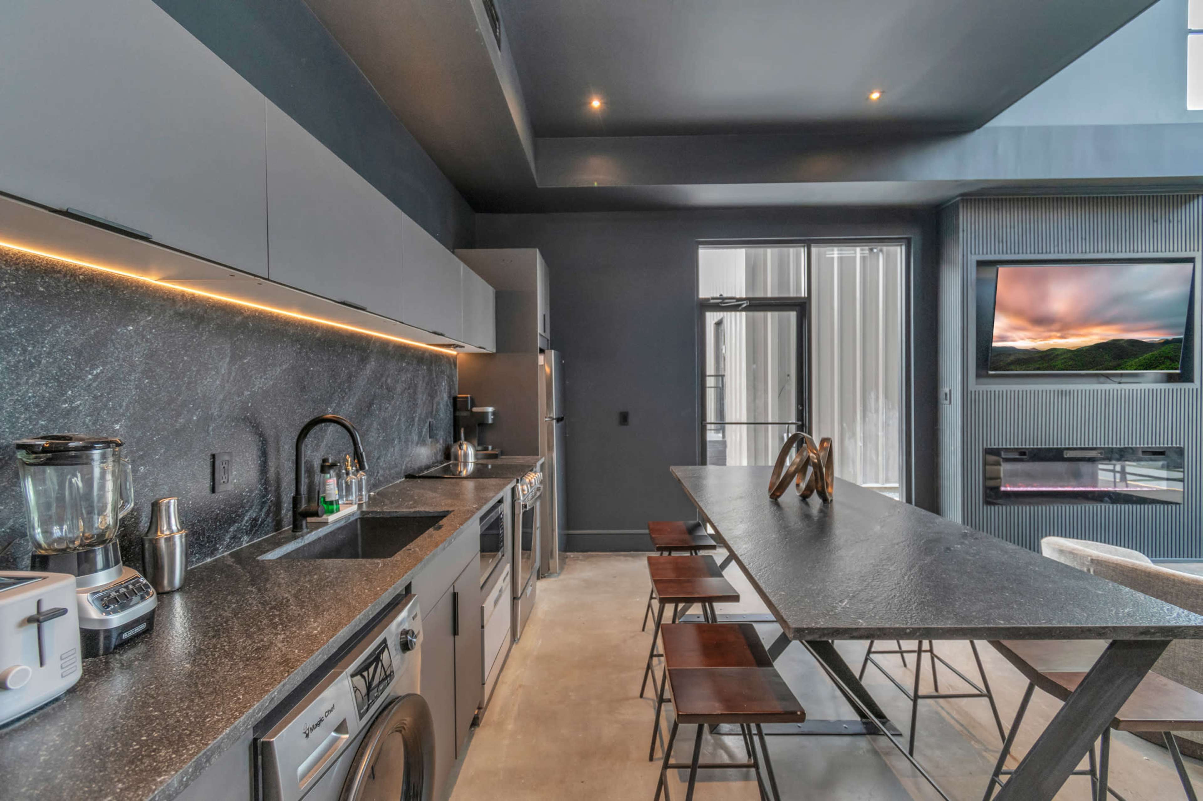 A modern kitchen features sleek gray cabinetry, a dark countertop, stainless steel appliances, and a minimalist dining table.