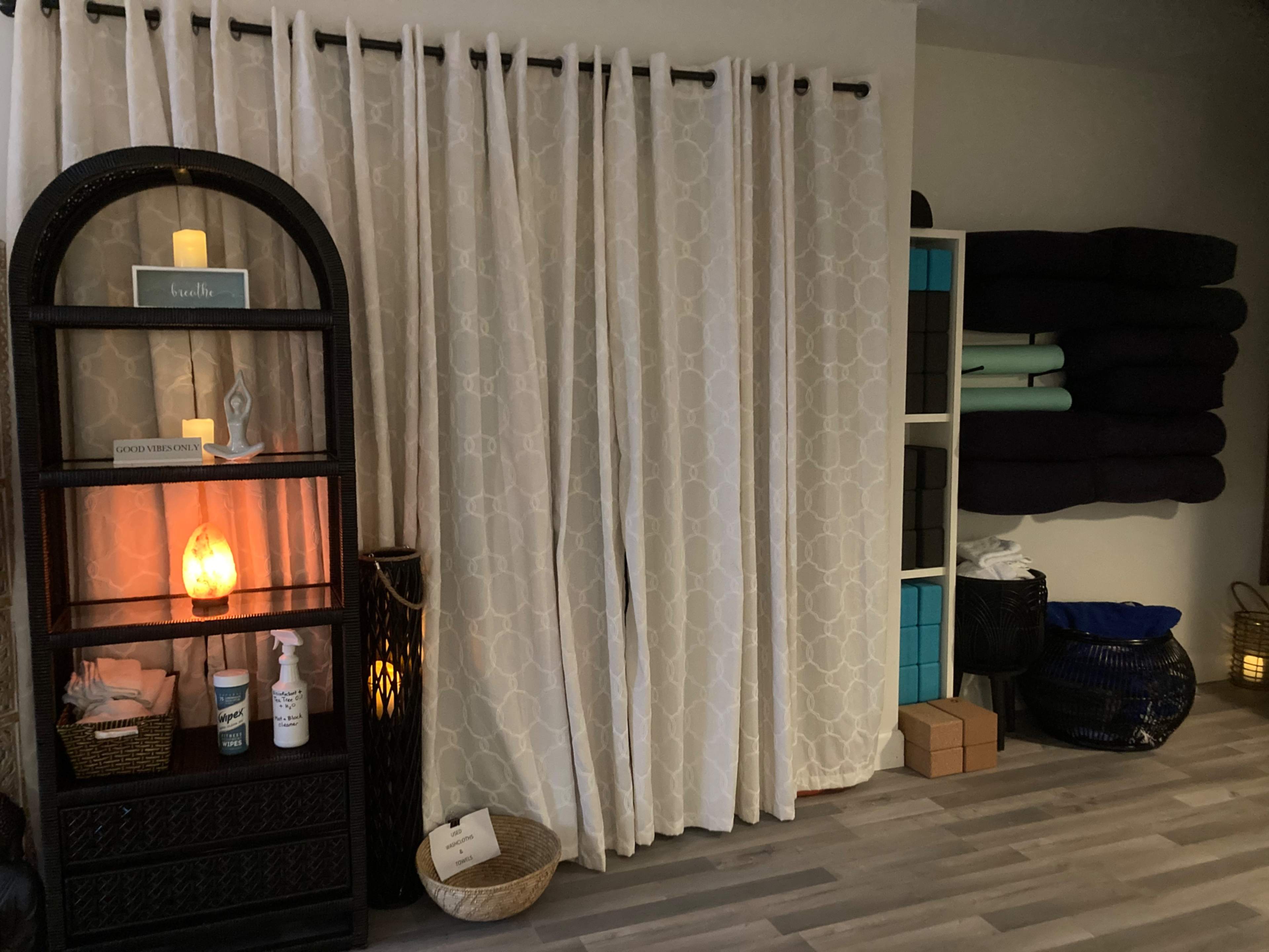 A serene interior space with curtains, a shelf displaying decorative items, a salt lamp, storage baskets, and neatly stacked mats.