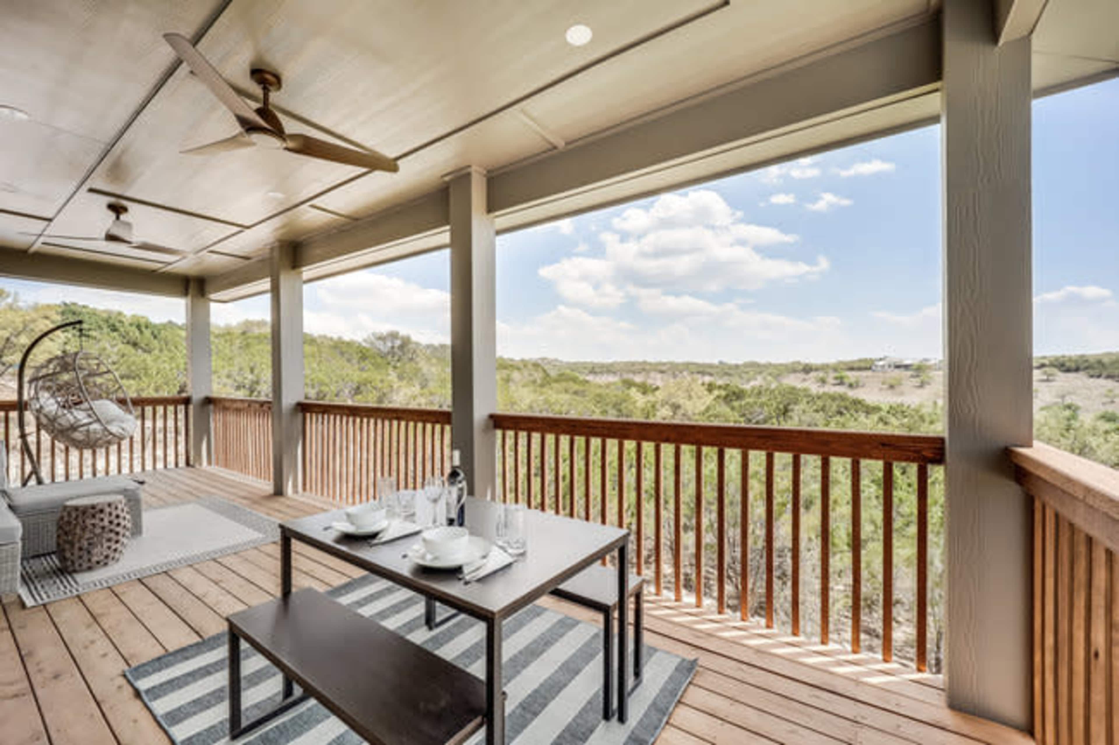 The image shows a spacious patio with a dining table, hanging chair, and scenic views of a landscape under a partly cloudy sky.