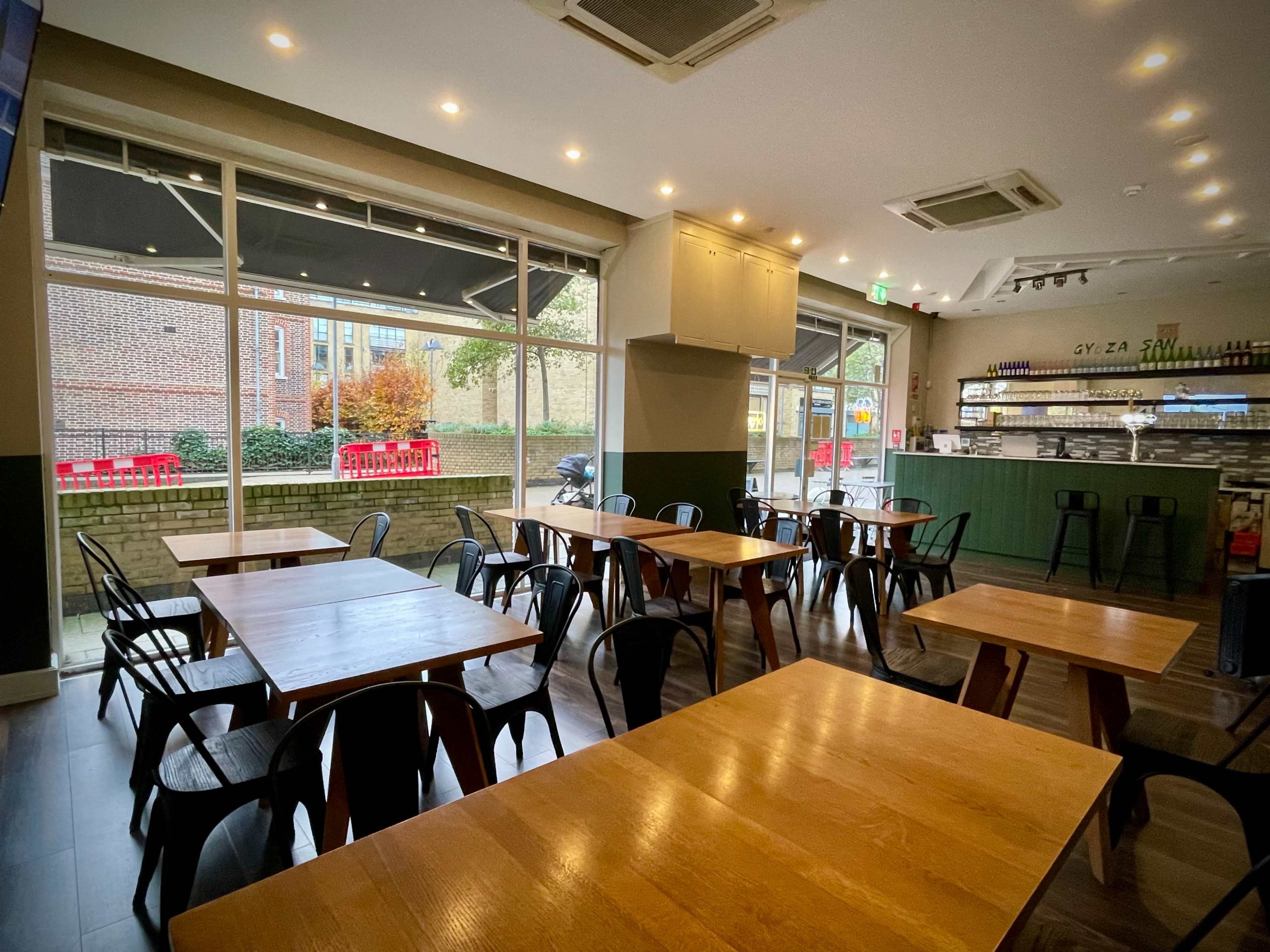 A modern restaurant interior with wooden tables and black metal chairs, large windows allowing natural light, and a bar area in the background.