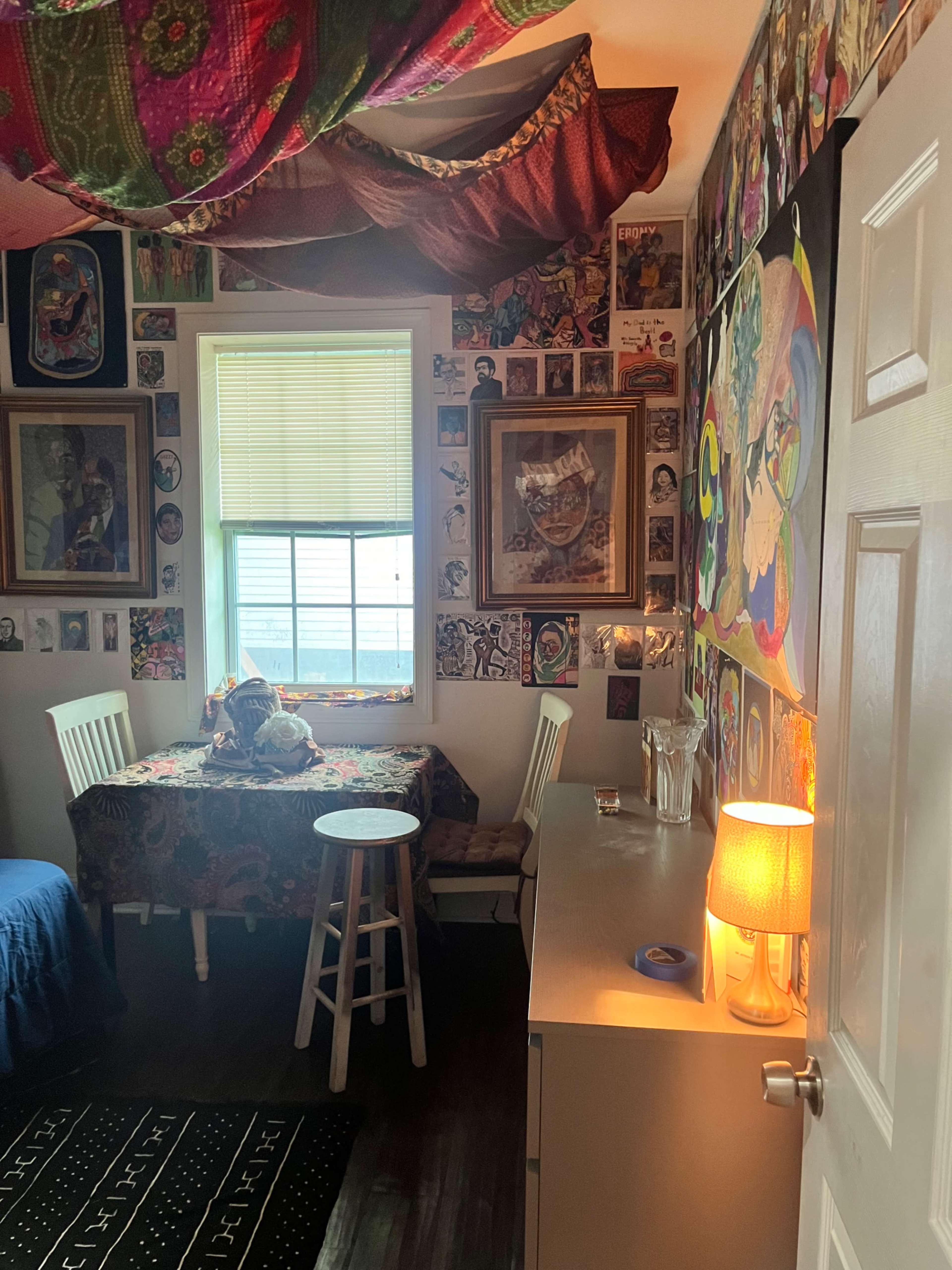 The image shows a small, decorated room with a vibrant ceiling drape, a table surrounded by chairs, and walls covered in various framed artworks and portraits.