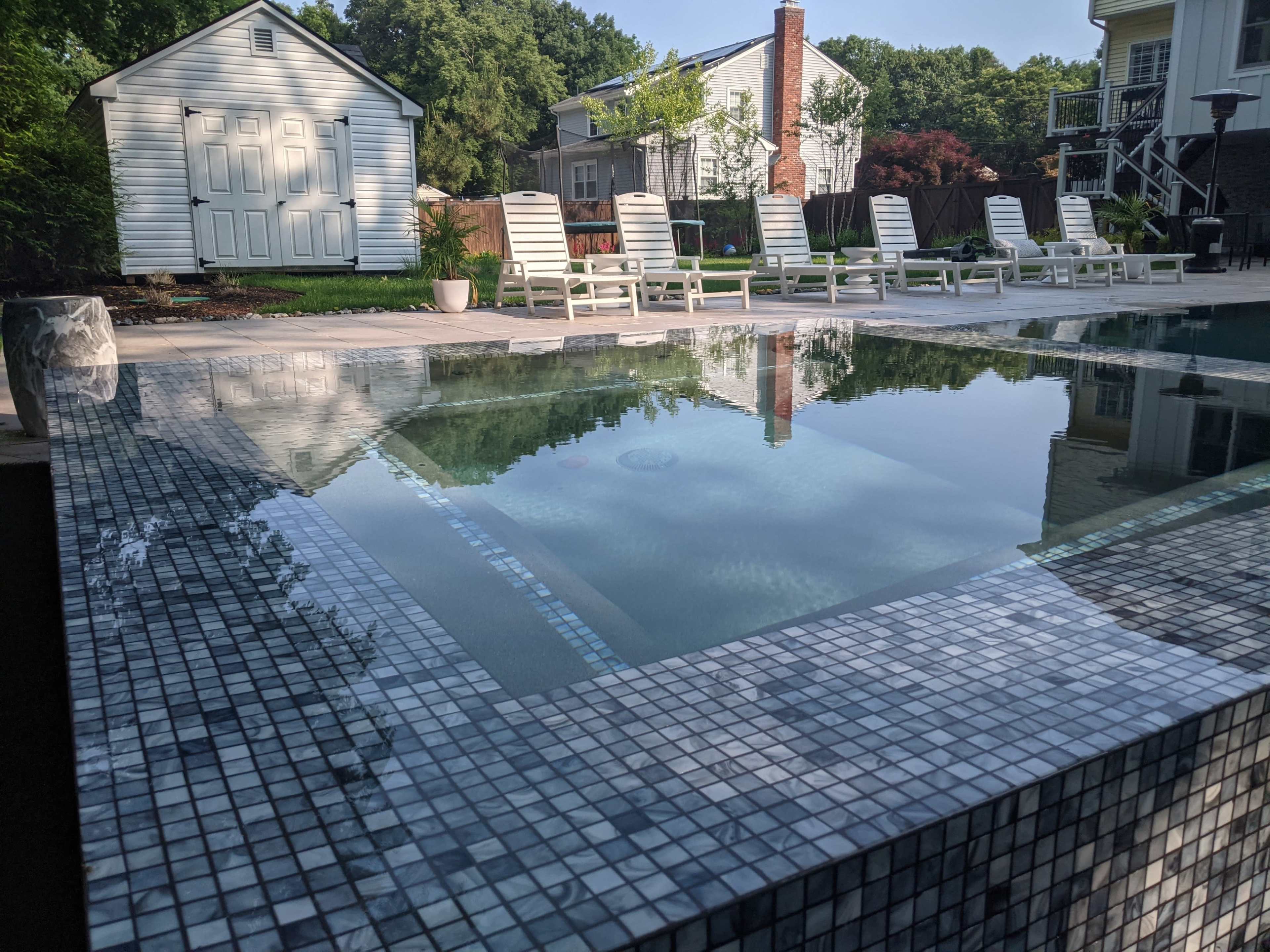 A modern swimming pool with mosaic tiles, surrounded by lounge chairs, and a white shed visible in the background.