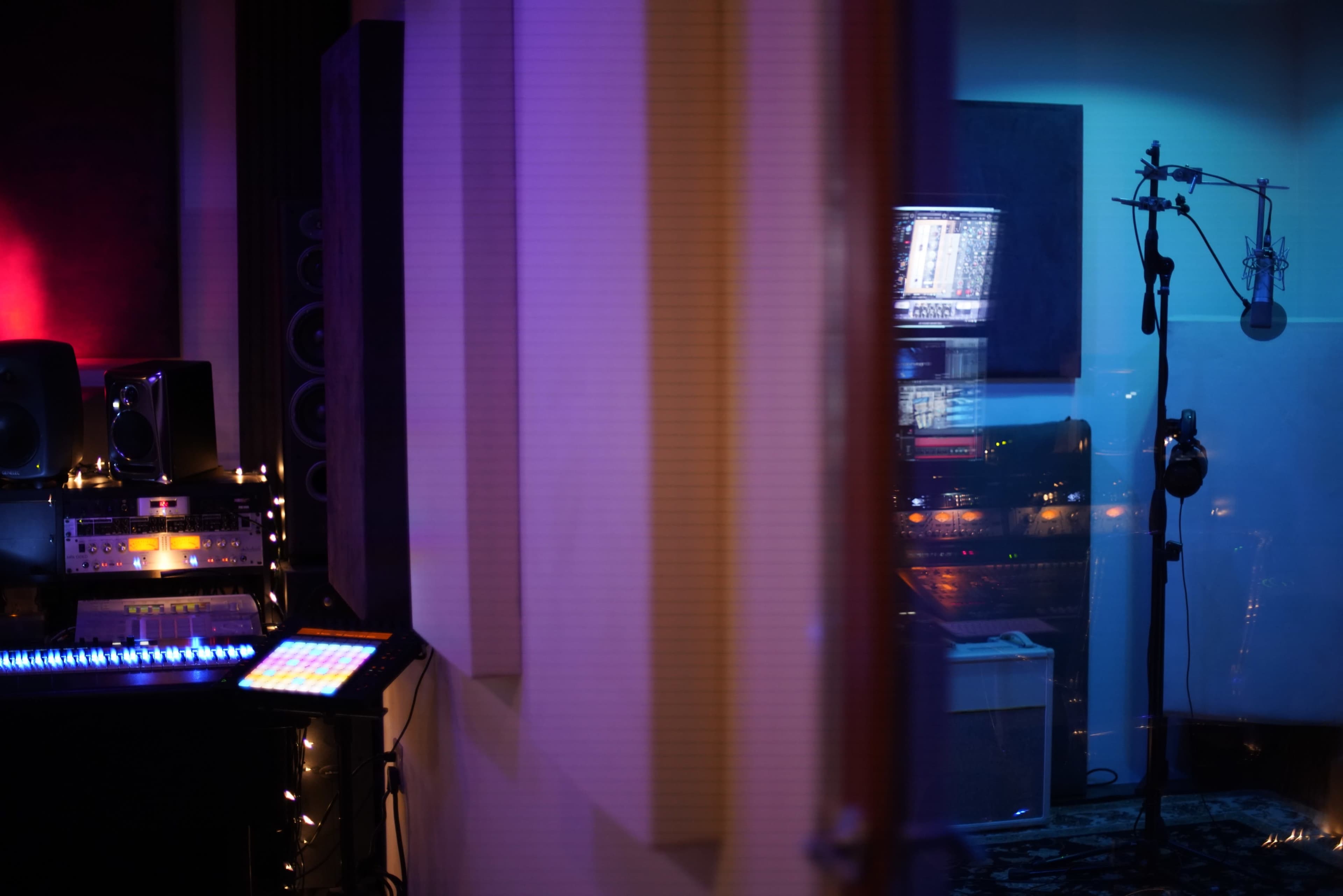 The image shows a music studio setup with sound equipment on the left and a microphone station visible through a glass partition on the right.