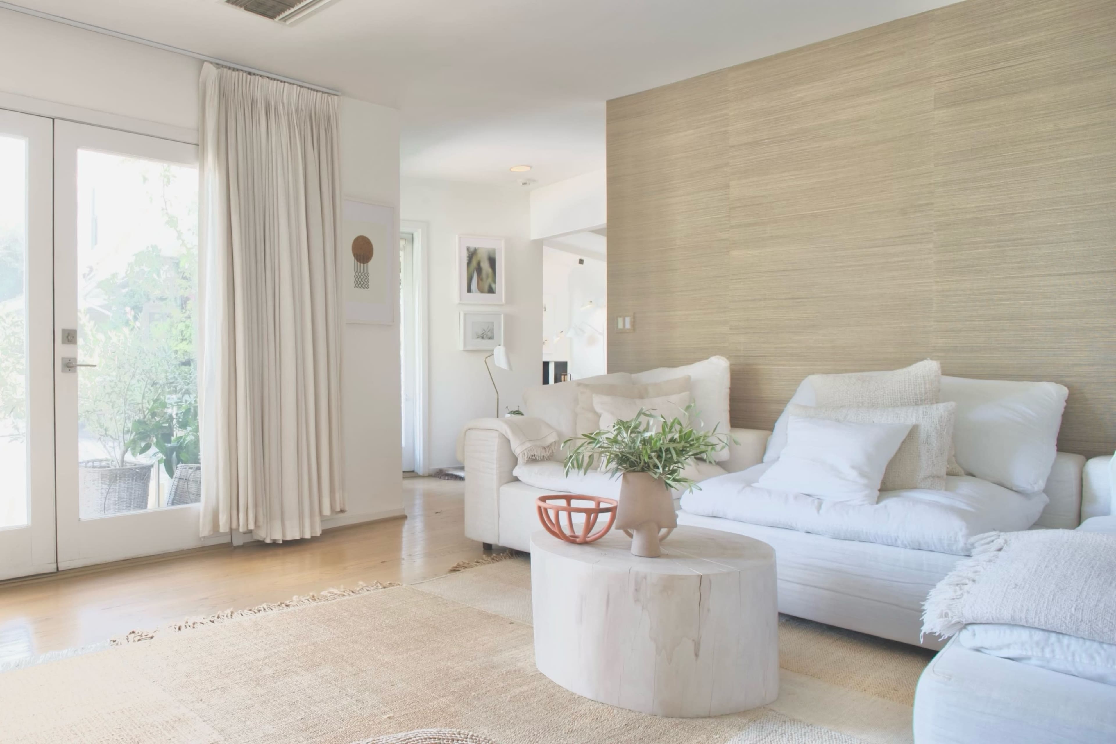 The image shows a modern living room with light-colored furniture, a round wooden coffee table, and large windows that let in natural light.