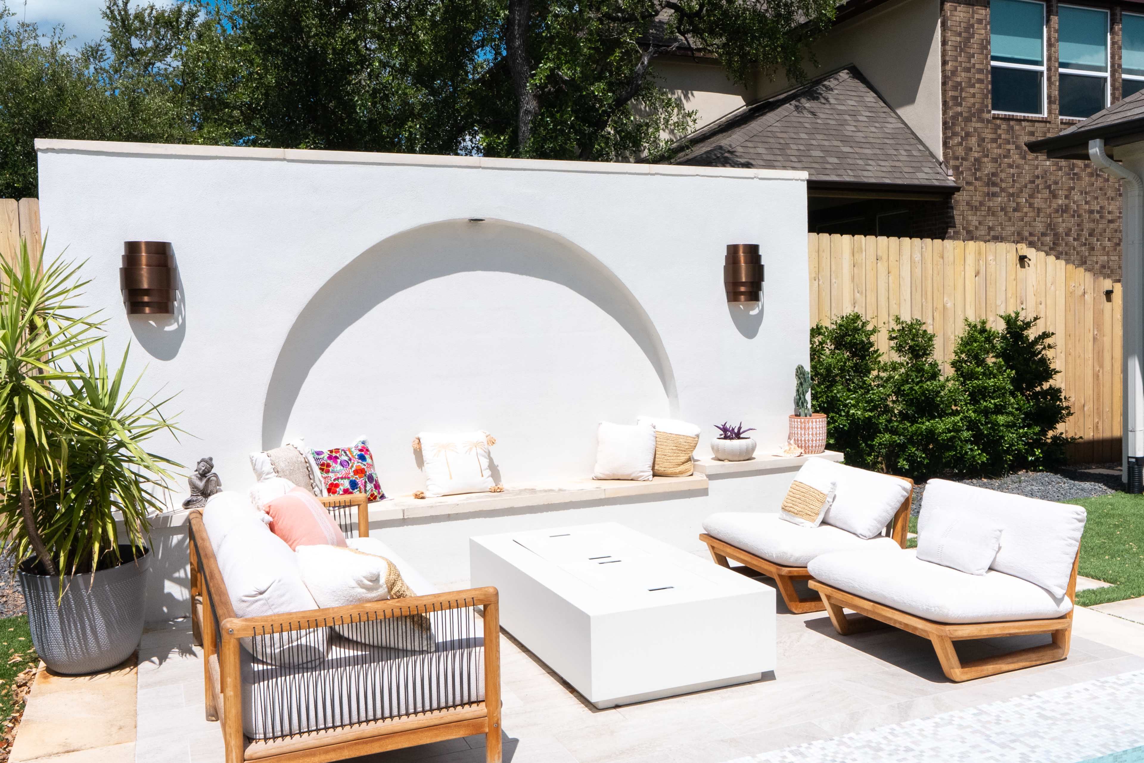 The image shows an outdoor seating area featuring modern furniture, a white wall with an arch design, and potted plants.
