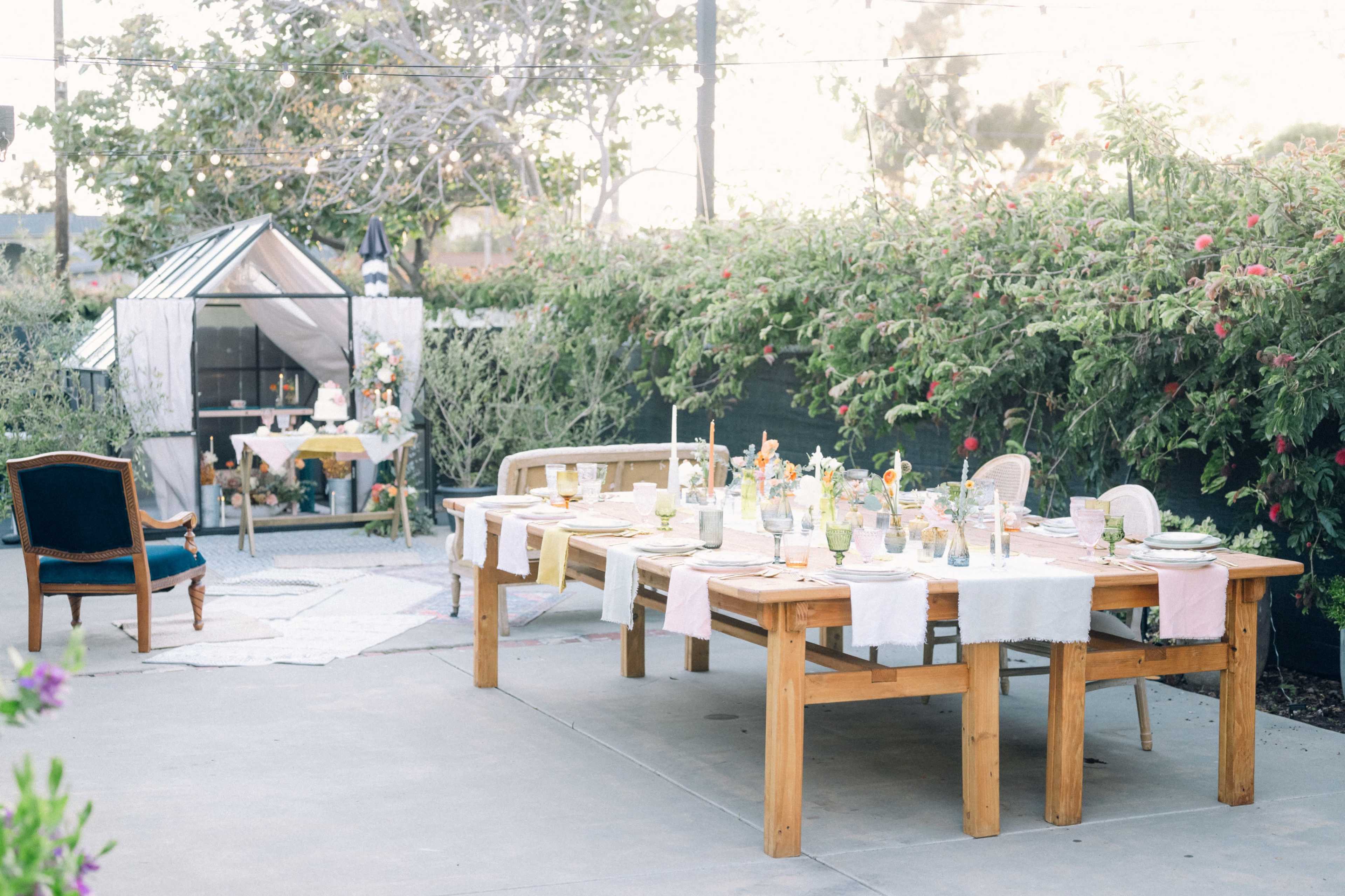 A long wooden table is set outdoors with a variety of dishes and glassware, surrounded by greenery and a small decorative greenhouse in the background.