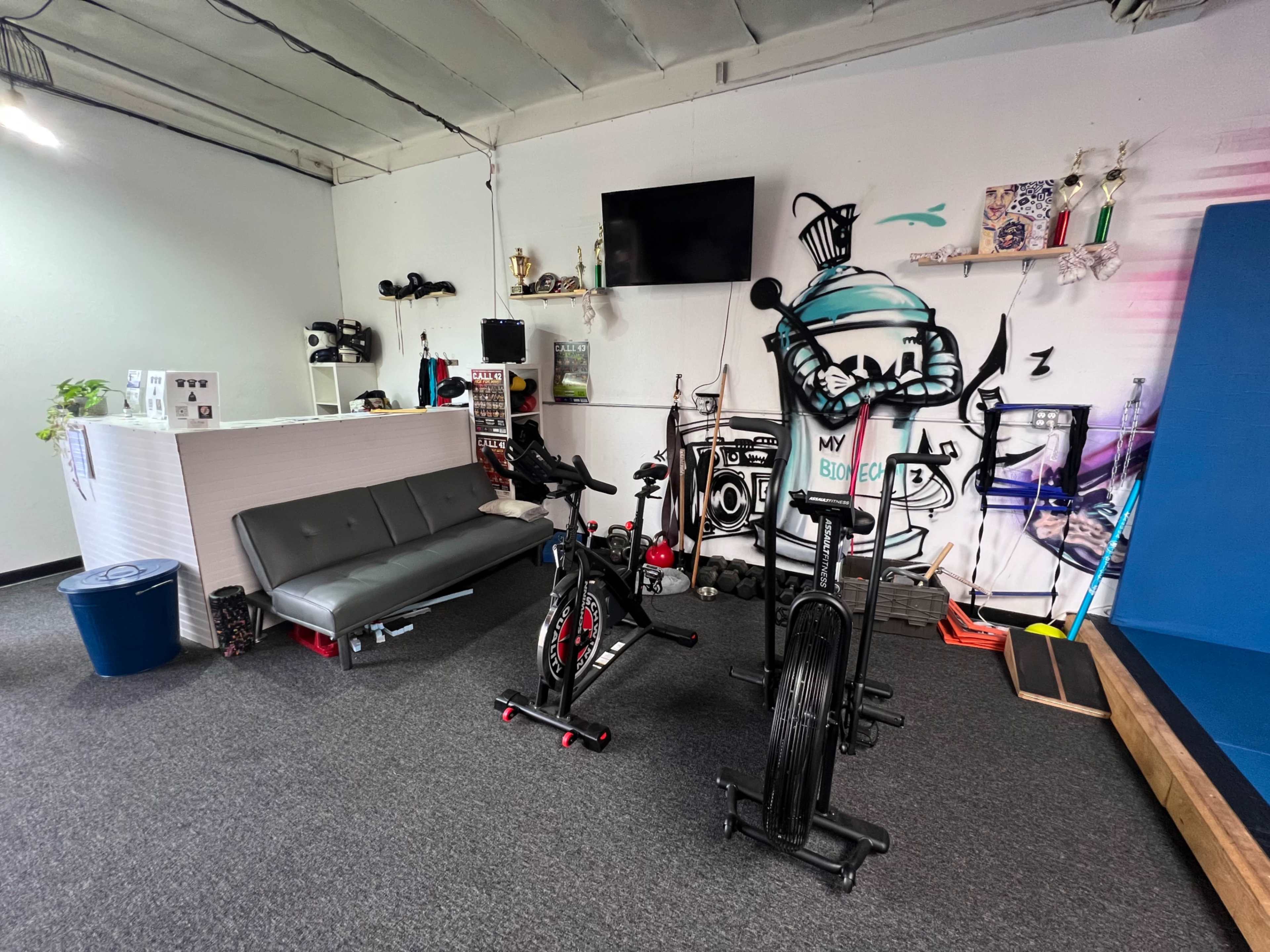 An indoor gym area featuring exercise bikes, a minimalist reception desk, and a graffiti mural on the wall.