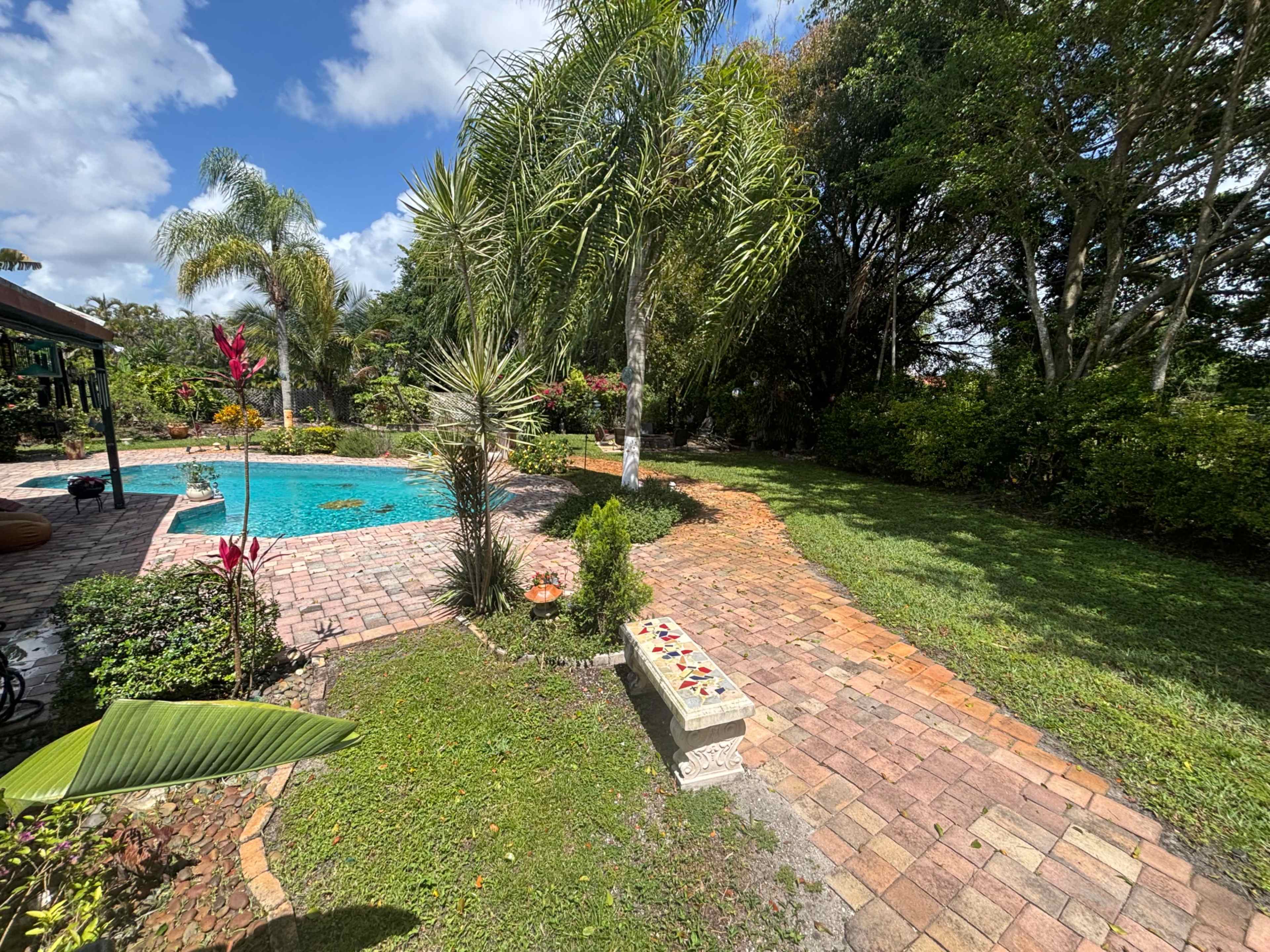 The image shows a landscaped backyard featuring a swimming pool surrounded by palm trees, flowers, and a brick pathway.
