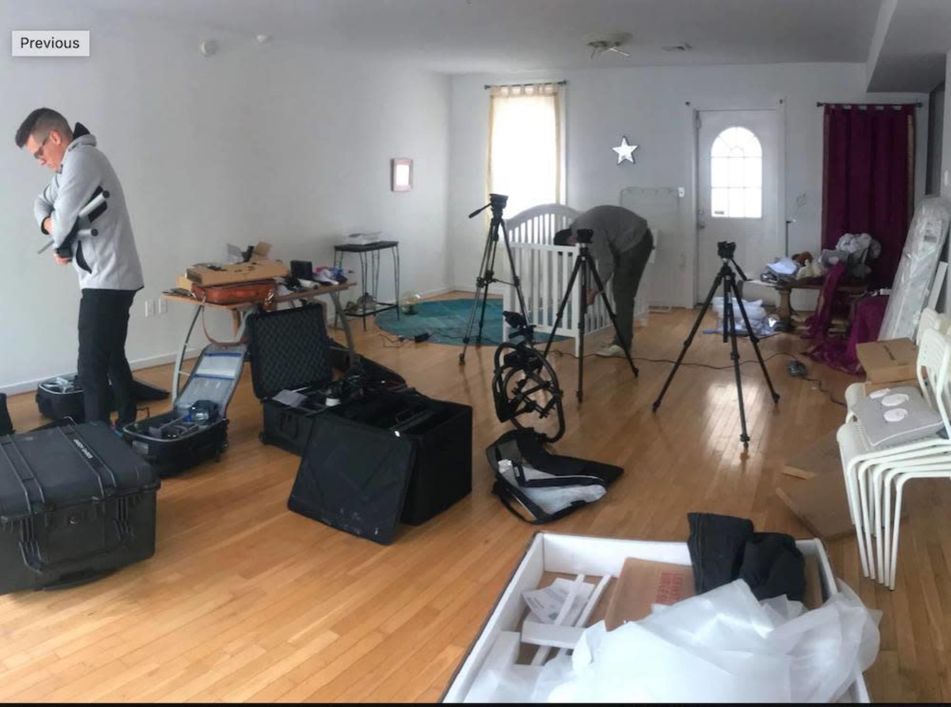 Two people are setting up equipment in a cluttered room with wooden floors and various items scattered around.