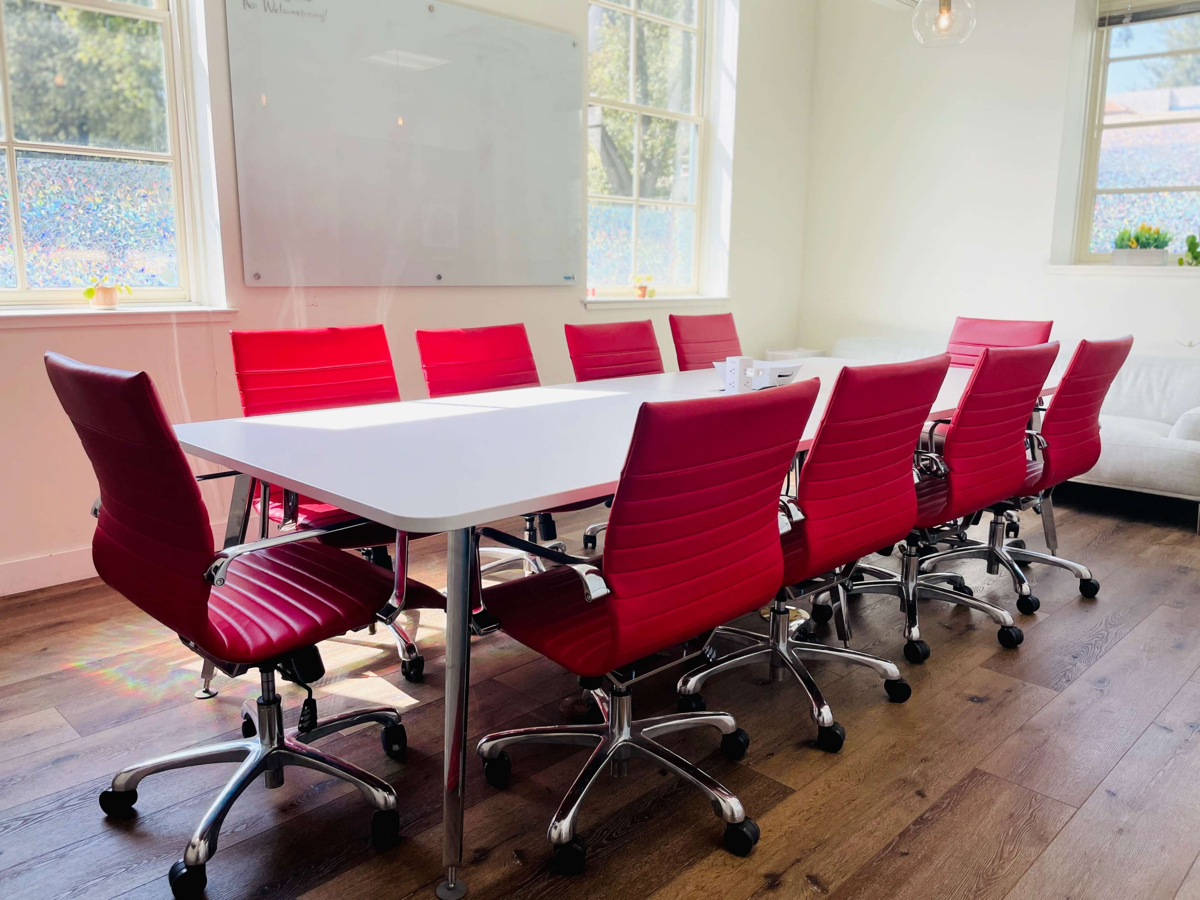 A spacious conference room features a long white table surrounded by eight red rolling chairs.