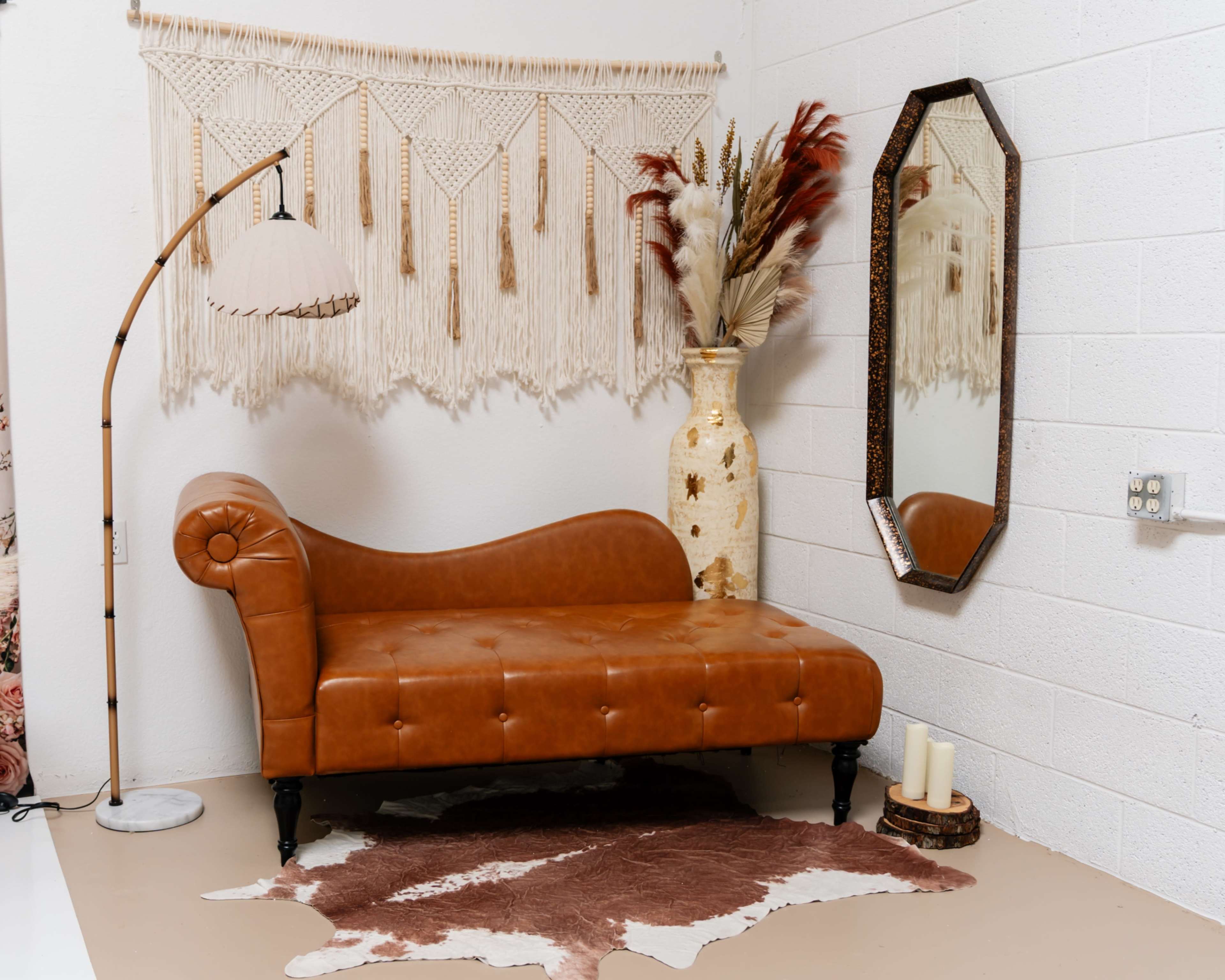 The image features a brown leather chaise lounge with a unique backrest positioned against a white wall adorned with a macramé wall hanging, complemented by a decorative mirror and a vase with dried plants.