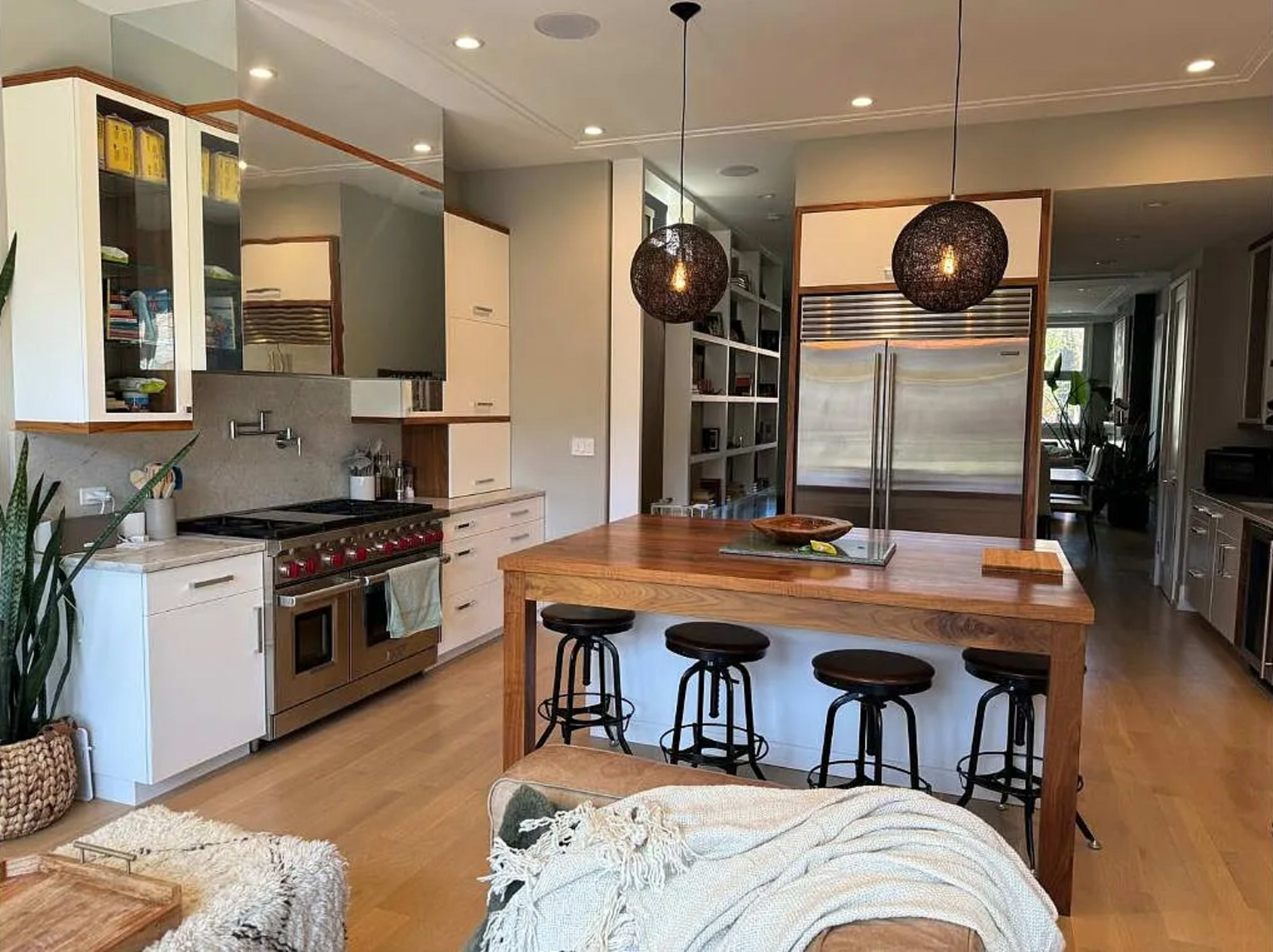 The image shows a modern kitchen with stainless steel appliances, a wooden island with four bar stools, and shelves lined with kitchenware and decor.