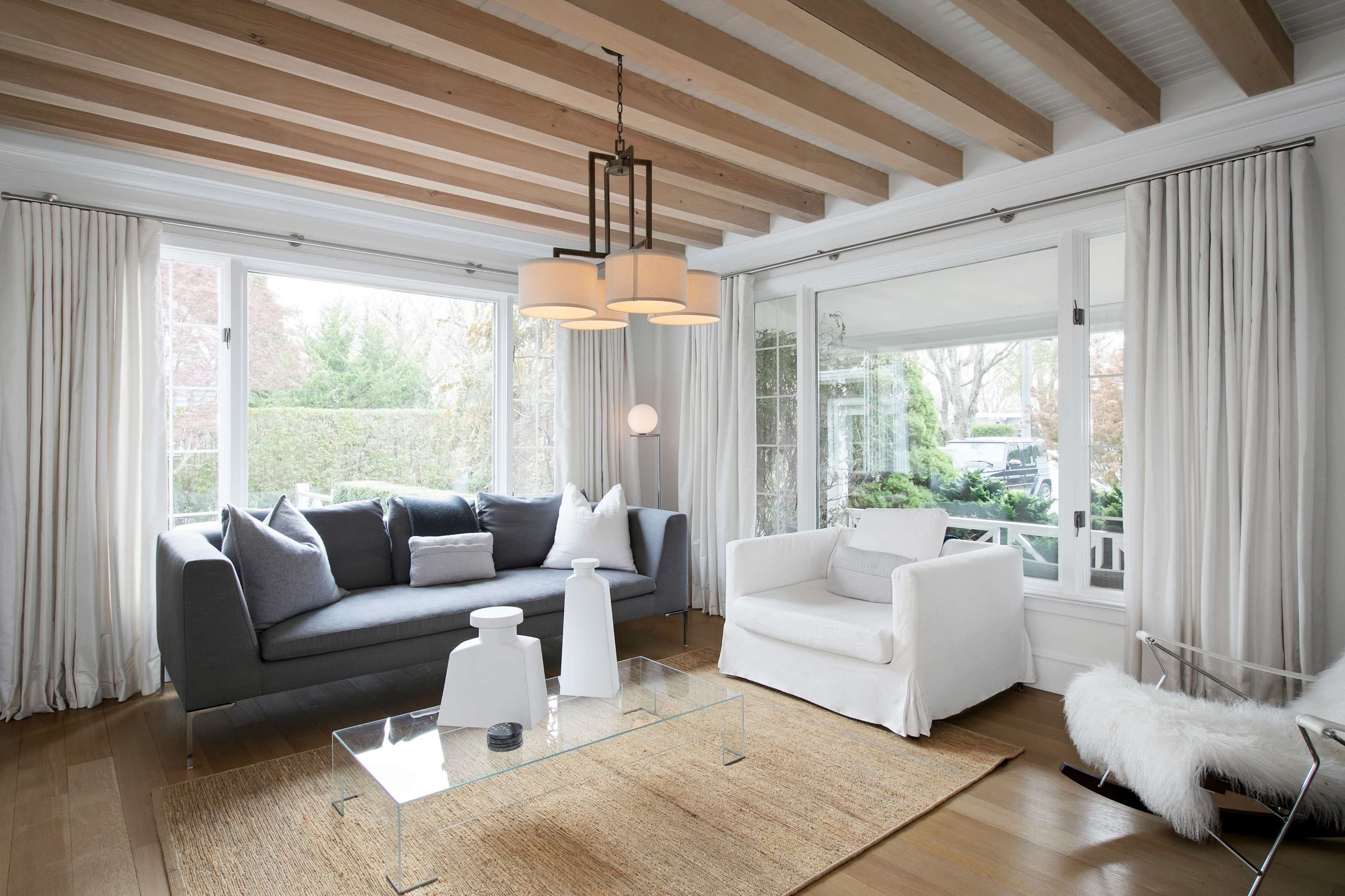 A living room features a gray sofa, a white armchair, and a glass coffee table on a woven rug, with large windows letting in natural light.