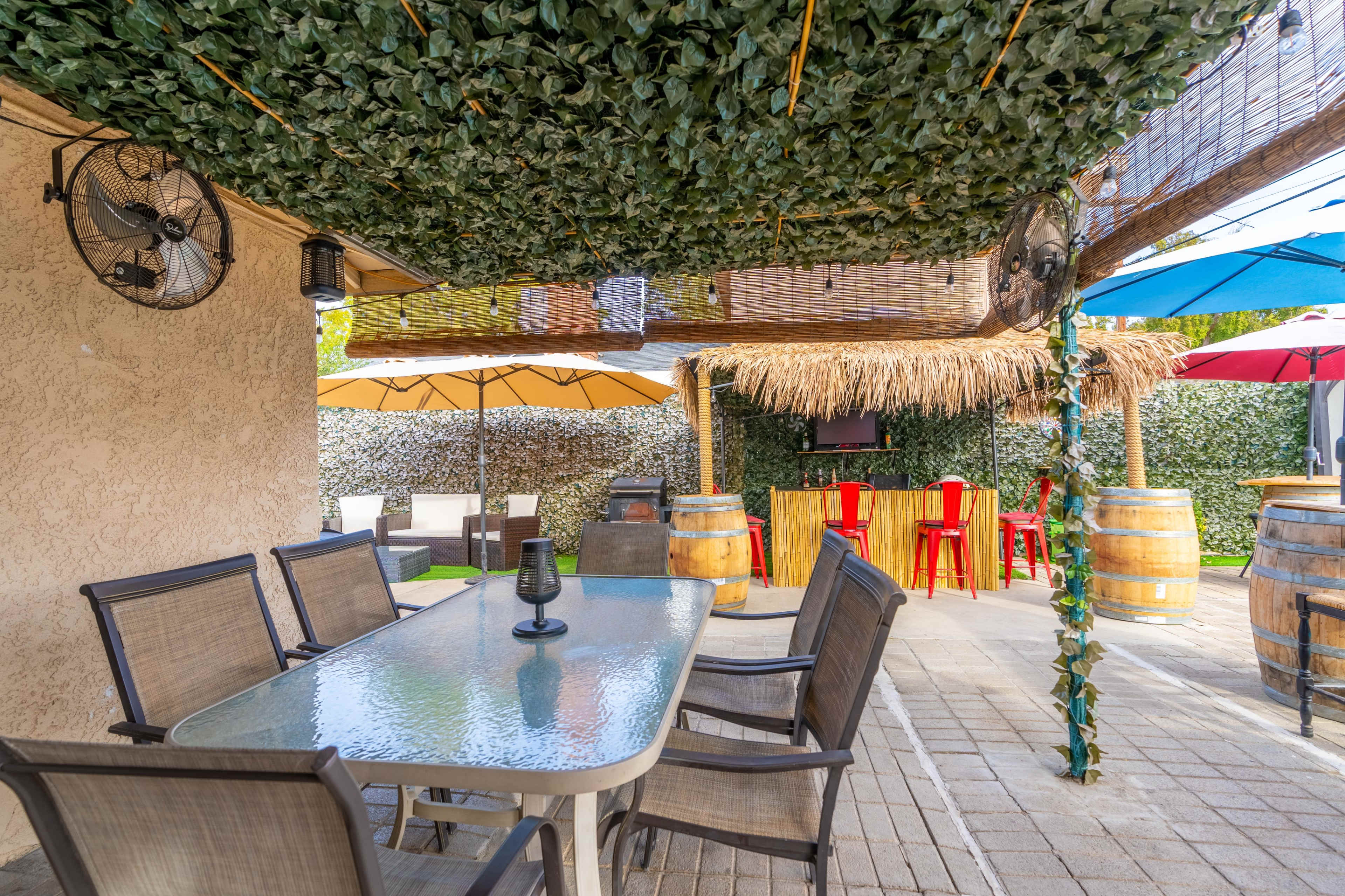 An outdoor patio area with a long table and chairs, shaded by a leafy canopy, surrounded by umbrellas and decorative elements like barrels and a thatched bar.