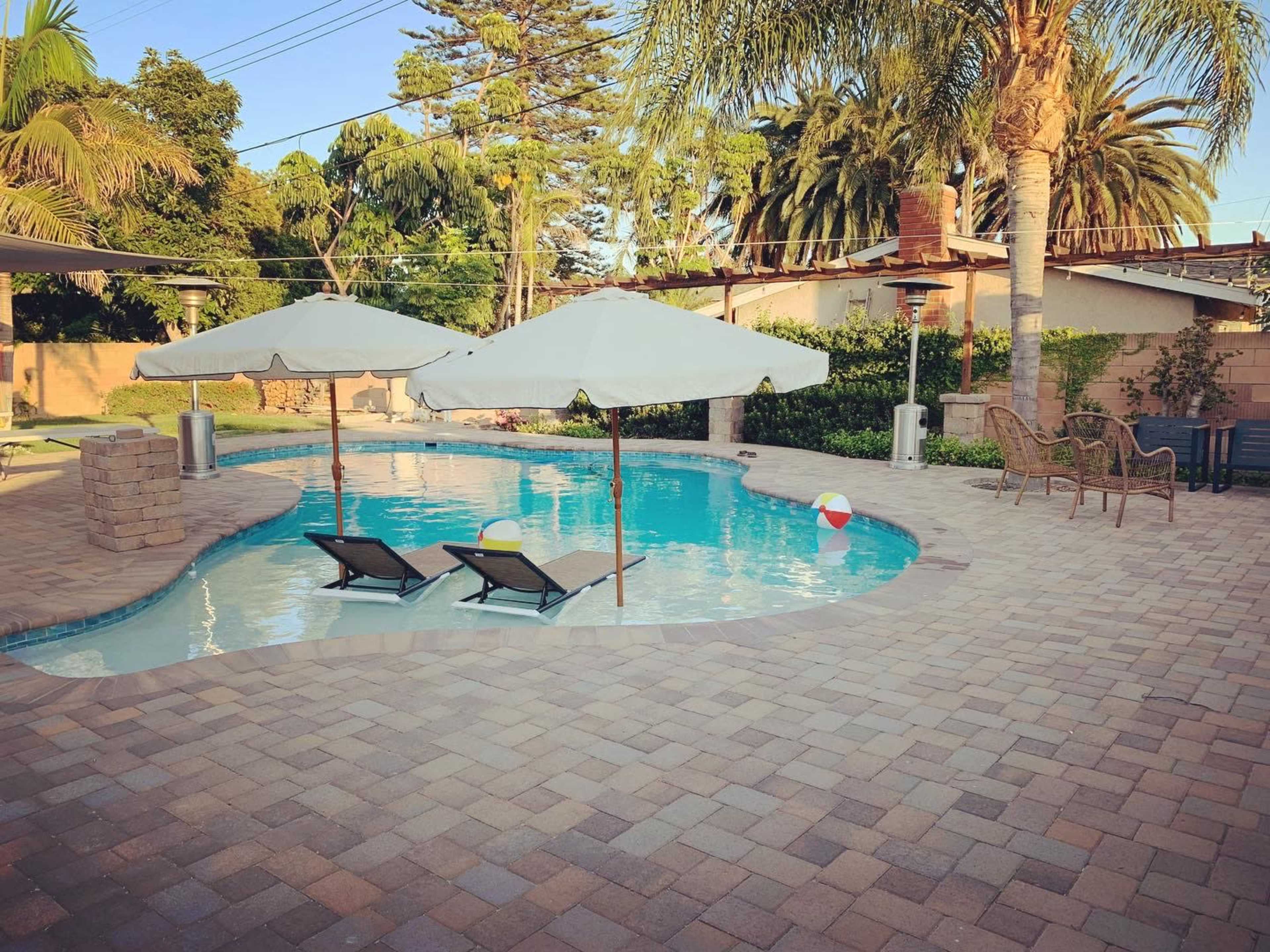 A pool area featuring two lounge chairs under umbrellas, surrounded by palm trees and a paved patio with a beach ball nearby.