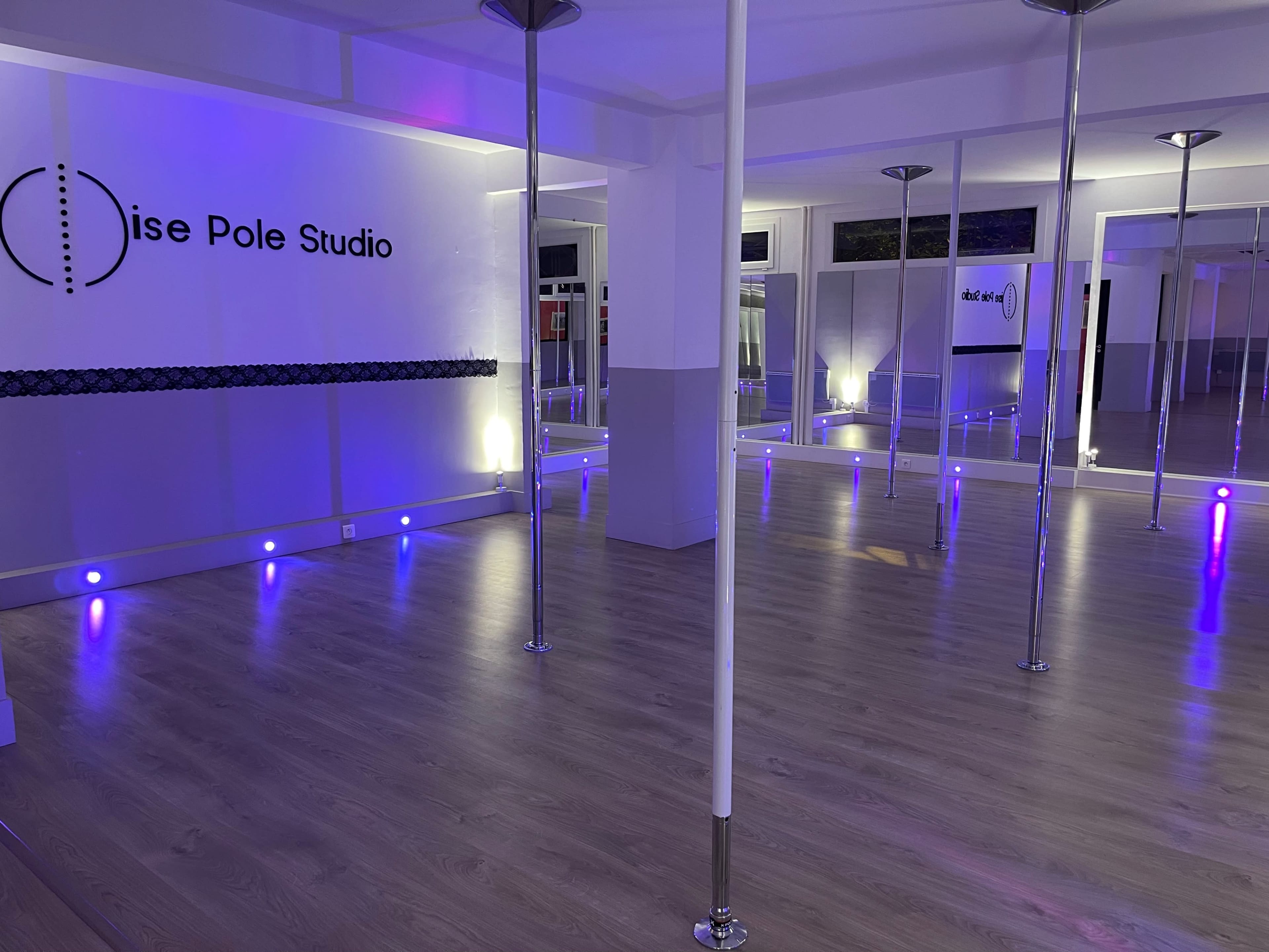 A spacious interior of a pole dance studio with polished wooden floors, several vertical poles, and soft purple lighting.