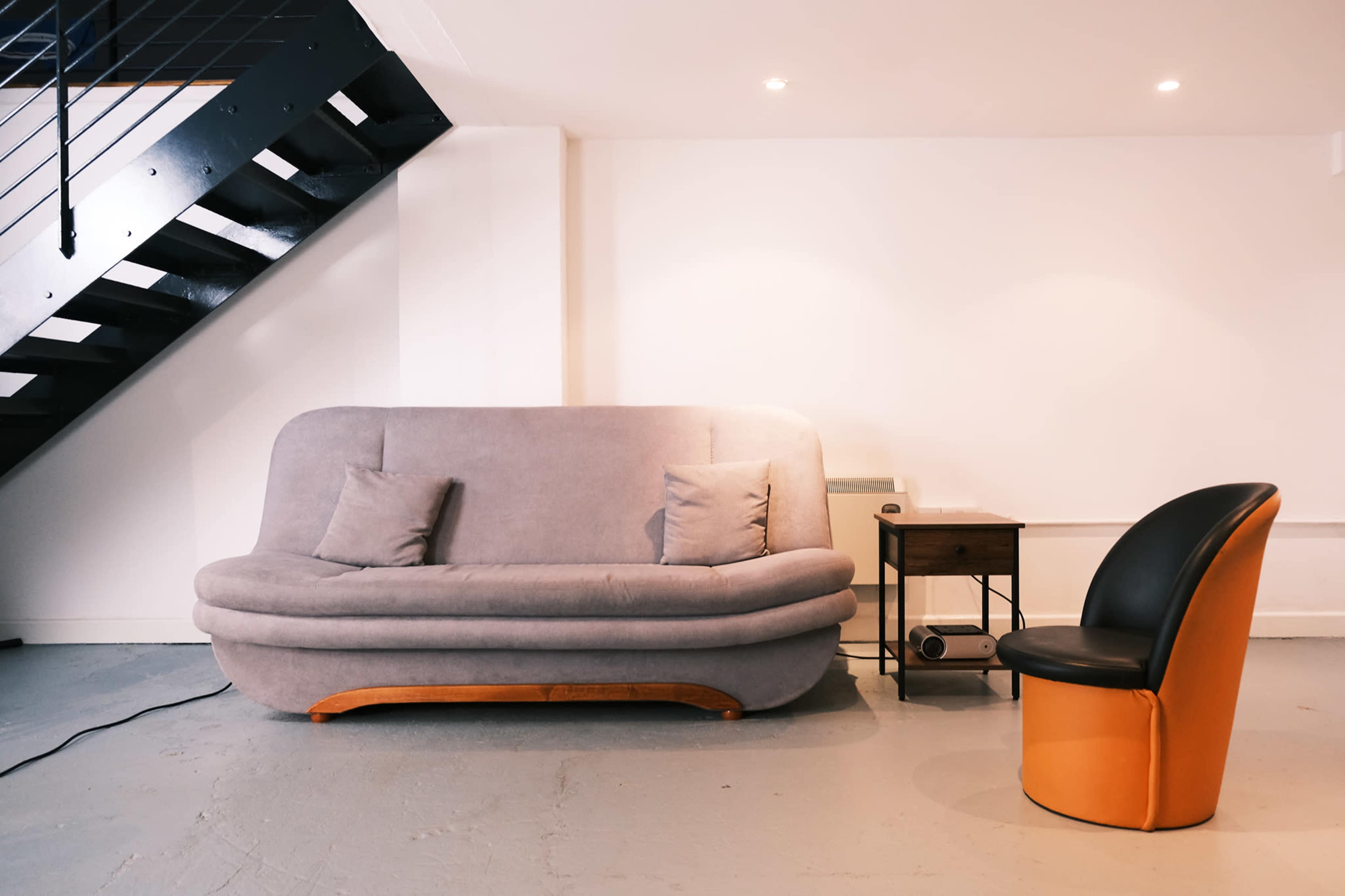 A gray sofa with two pillows is placed next to a wooden side table and a black and orange chair, set against a simple white wall and staircase.