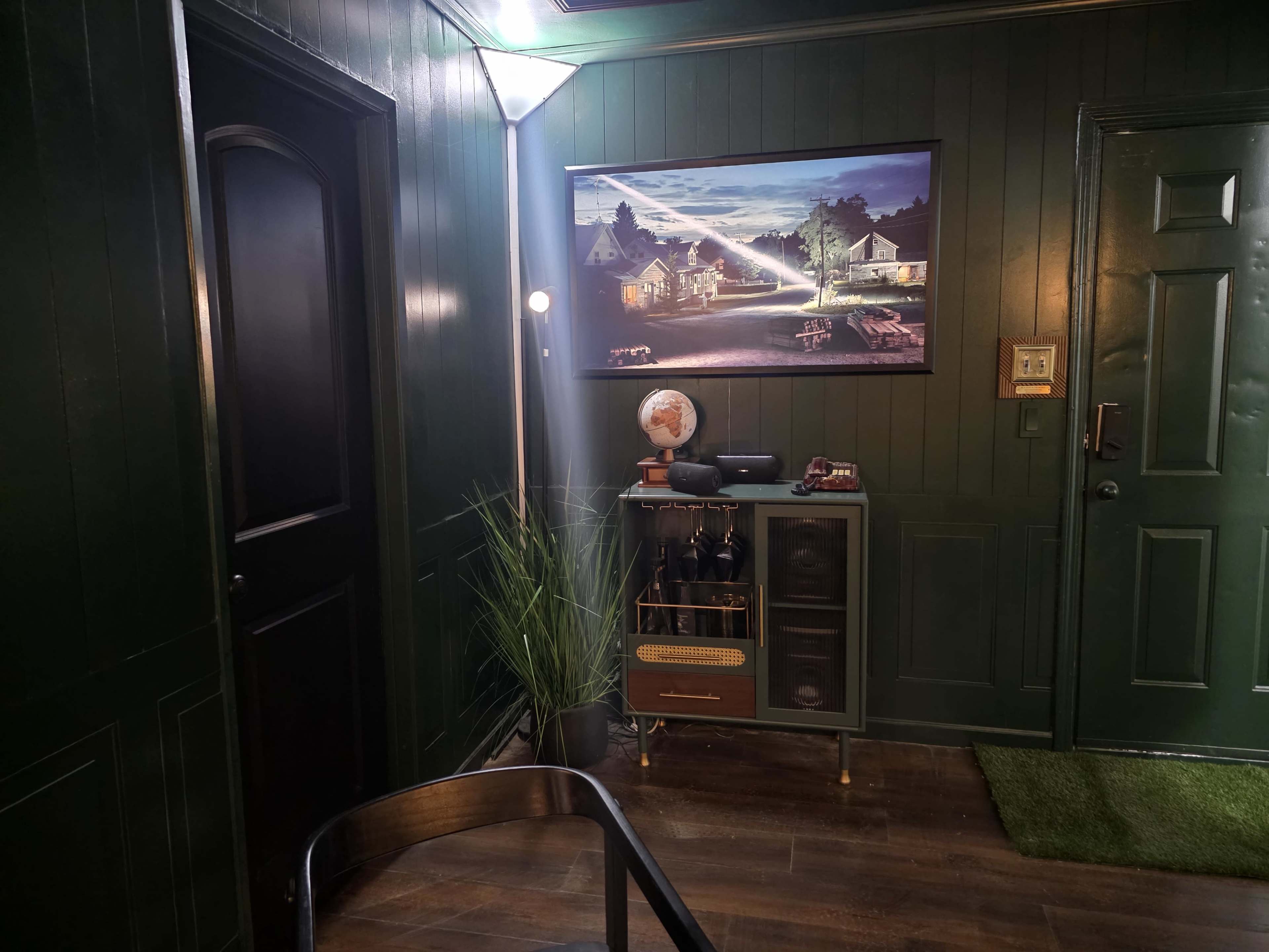 The scene features a dark green room with a door, a wall-mounted painting of a nighttime landscape, and a small cabinet with decorative items and plants.