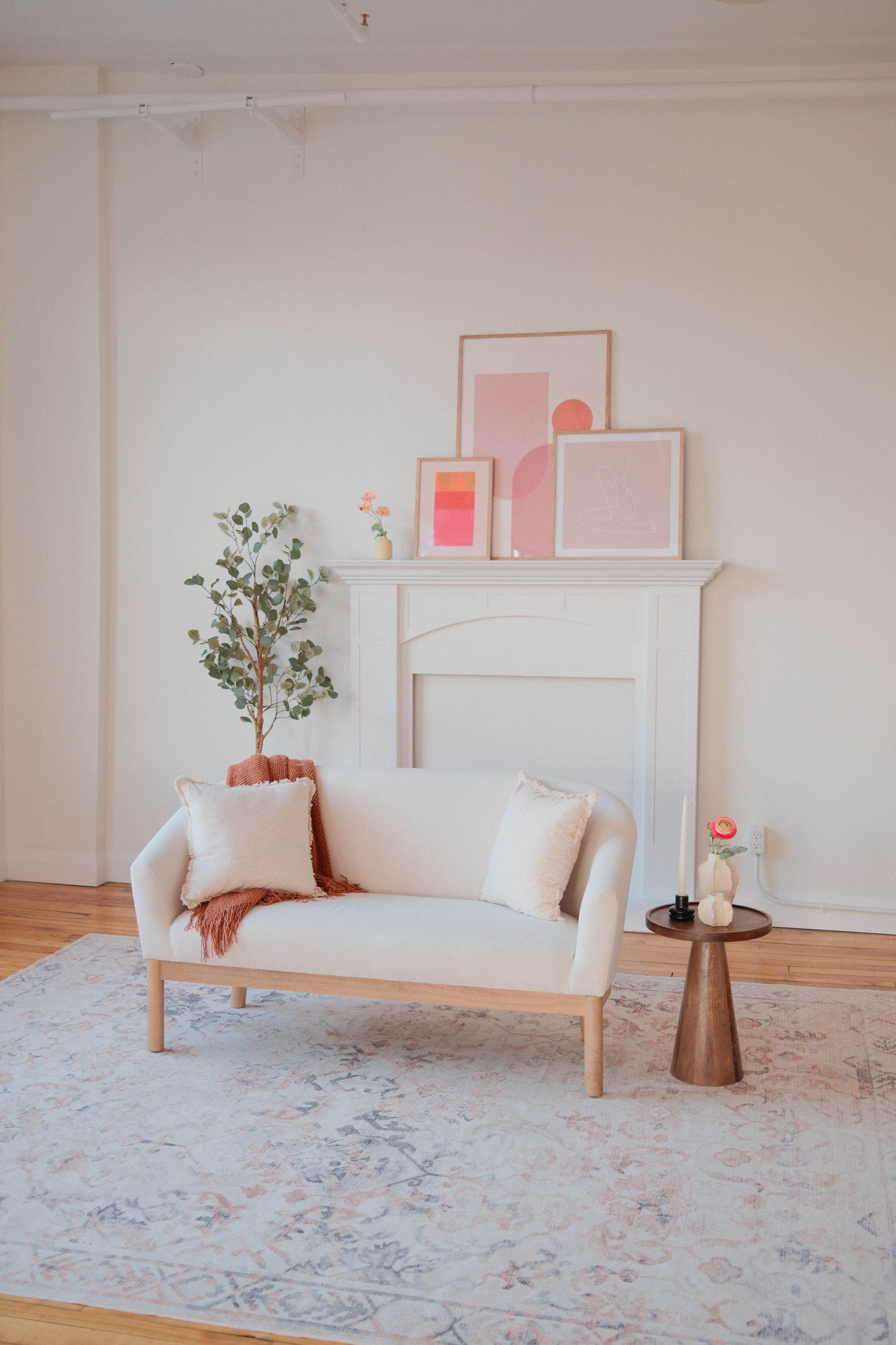 A light-colored sofa with decorative pillows is positioned near a mantel adorned with framed artwork, accompanied by a small plant and a side table on a patterned rug.