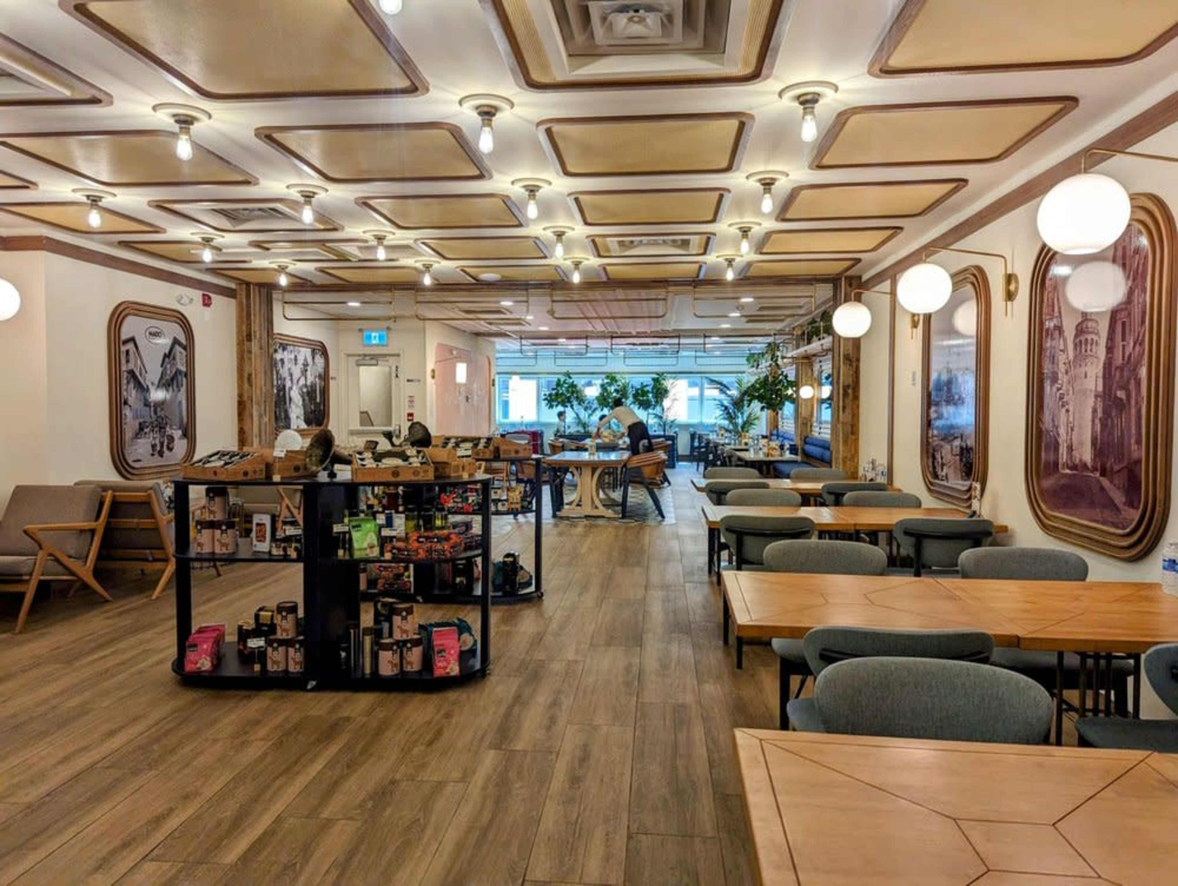 The image depicts a modern café interior with wooden flooring, small tables, and decorative lighting, surrounded by plants and a display shelf in the center.