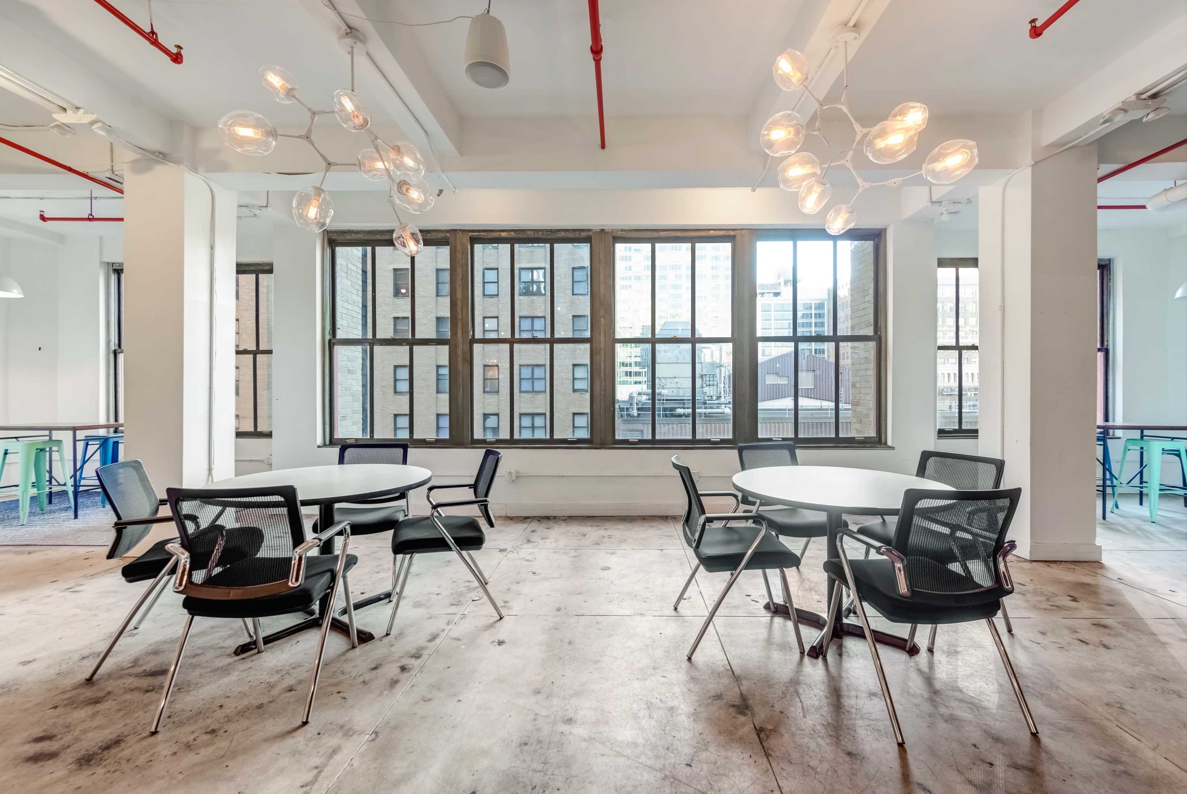 A modern office space with several round tables and chairs, large windows providing a view of a city, and distinctive light fixtures hanging from the ceiling.